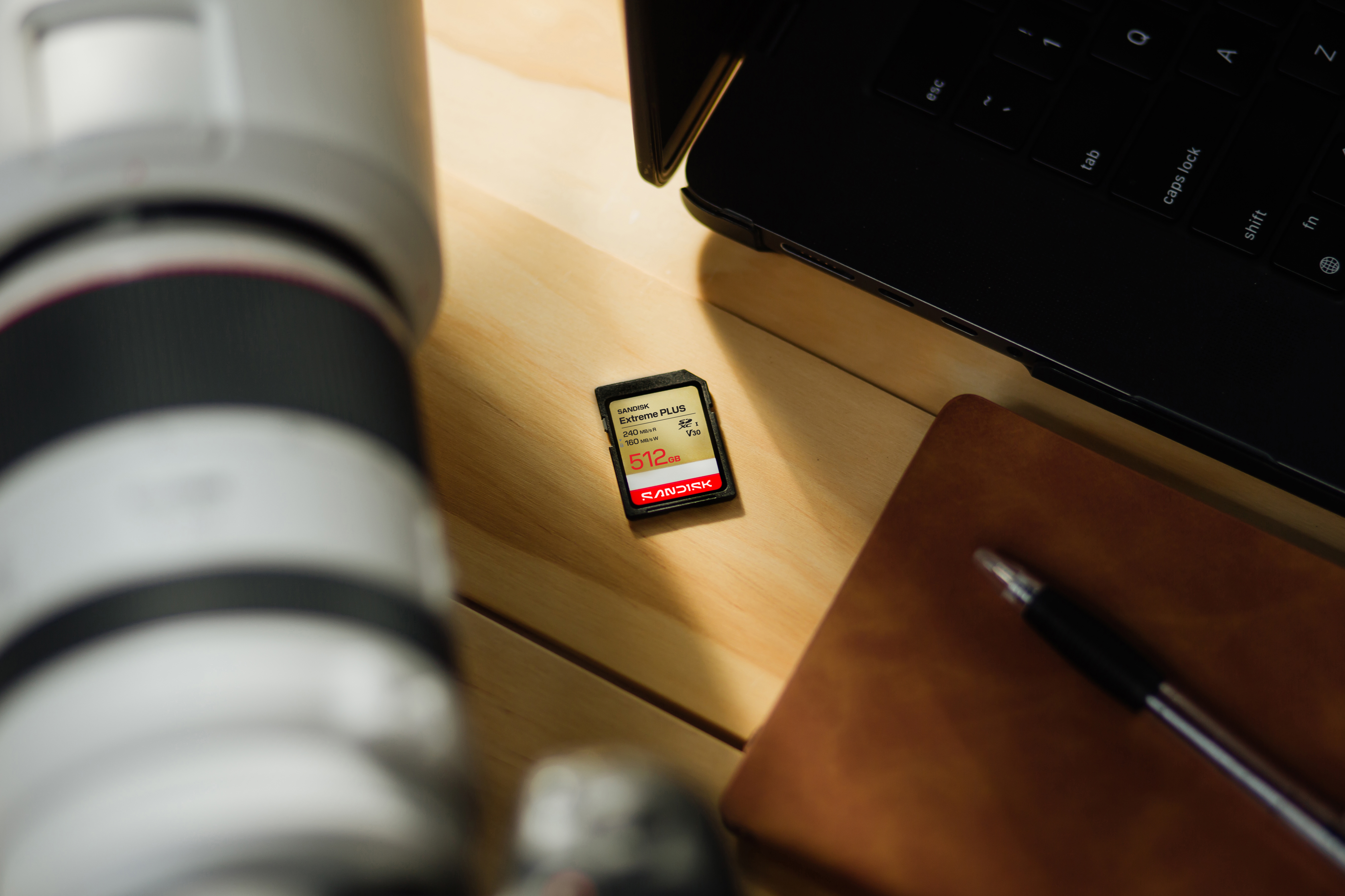 Ein SanDisk 512GB SD-Karte liegt neben einem Laptop und einer Kamera auf einem Holztisch.