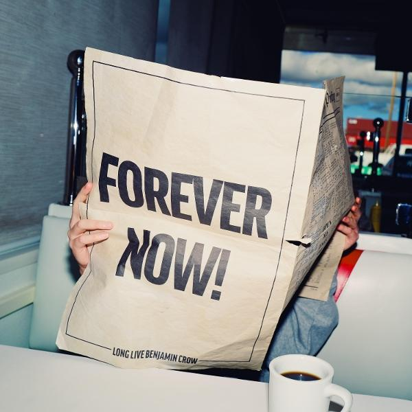 Jemand liest eine Zeitung mit "FOREVER NOW!". Eine Kaffeetasse steht daneben.