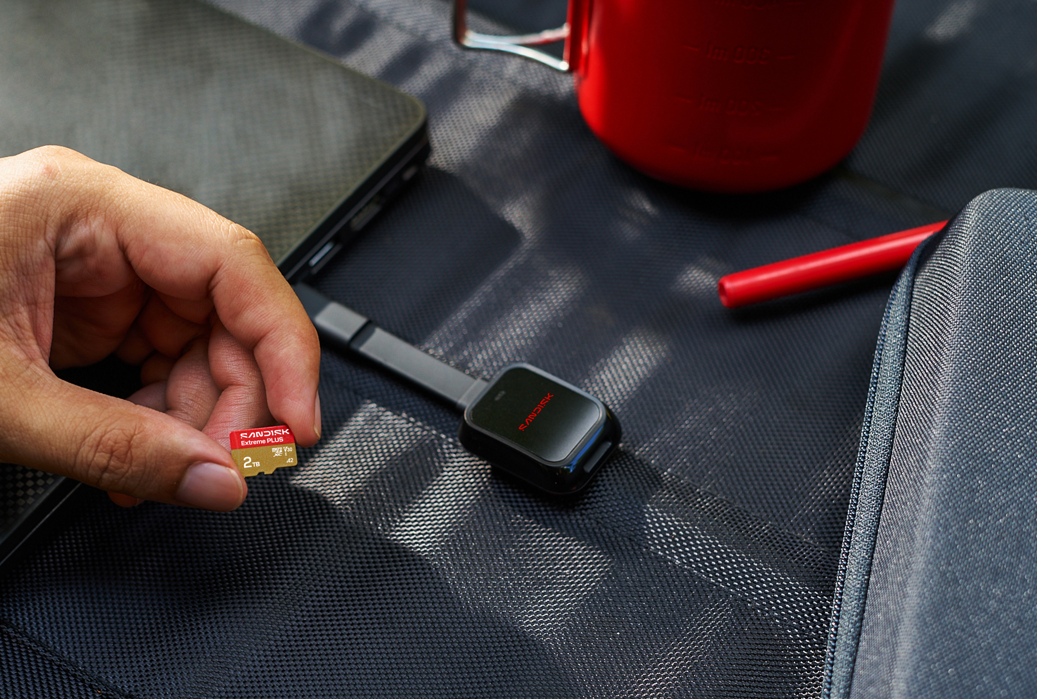 Eine Hand hält eine rote SanDisk 2TB MicroSD-Karte neben einem schwarzen Lesegerät.
