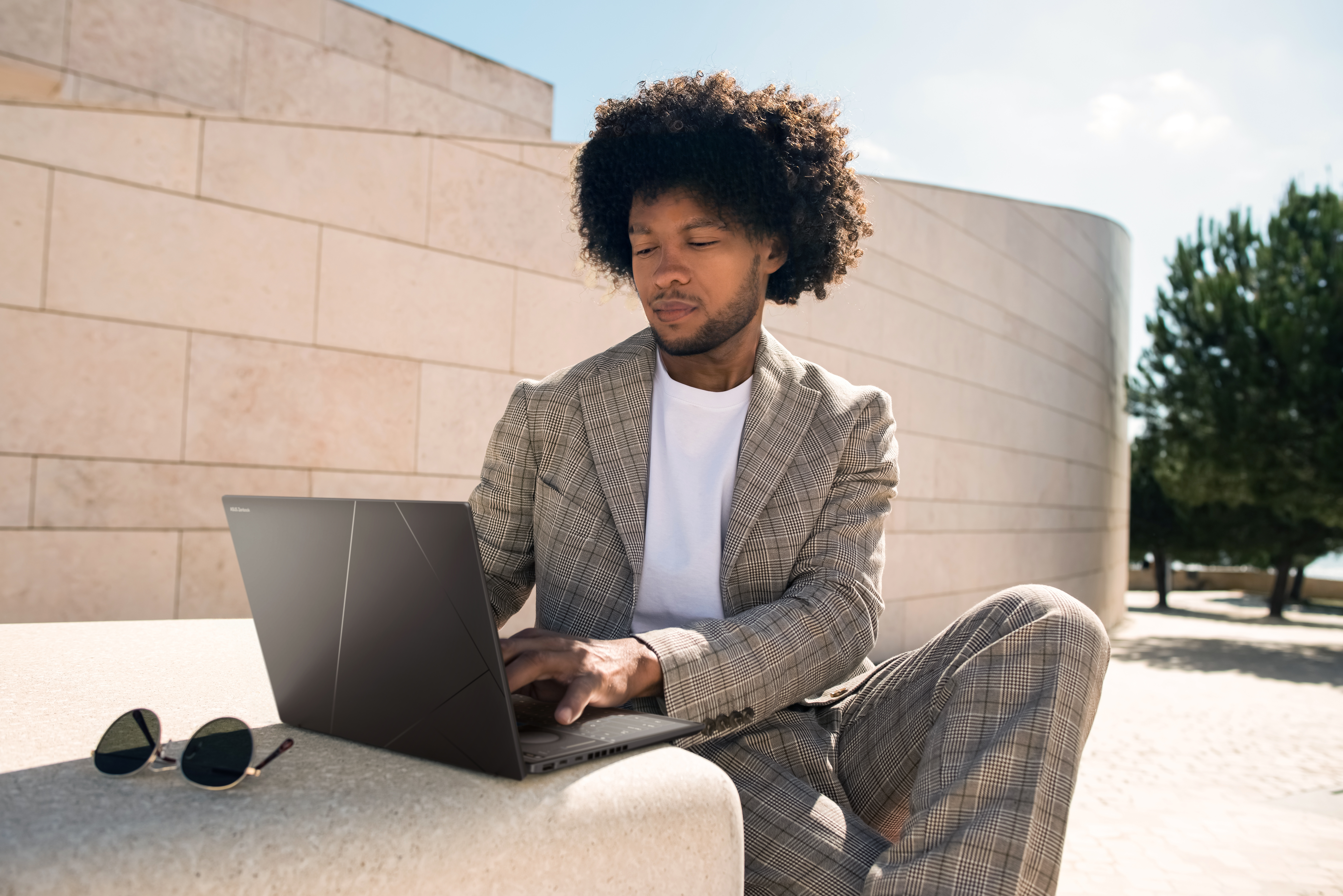 Mann mit Afro sitzt draußen und arbeitet an einem Laptop. Sonnenbrille liegt daneben.