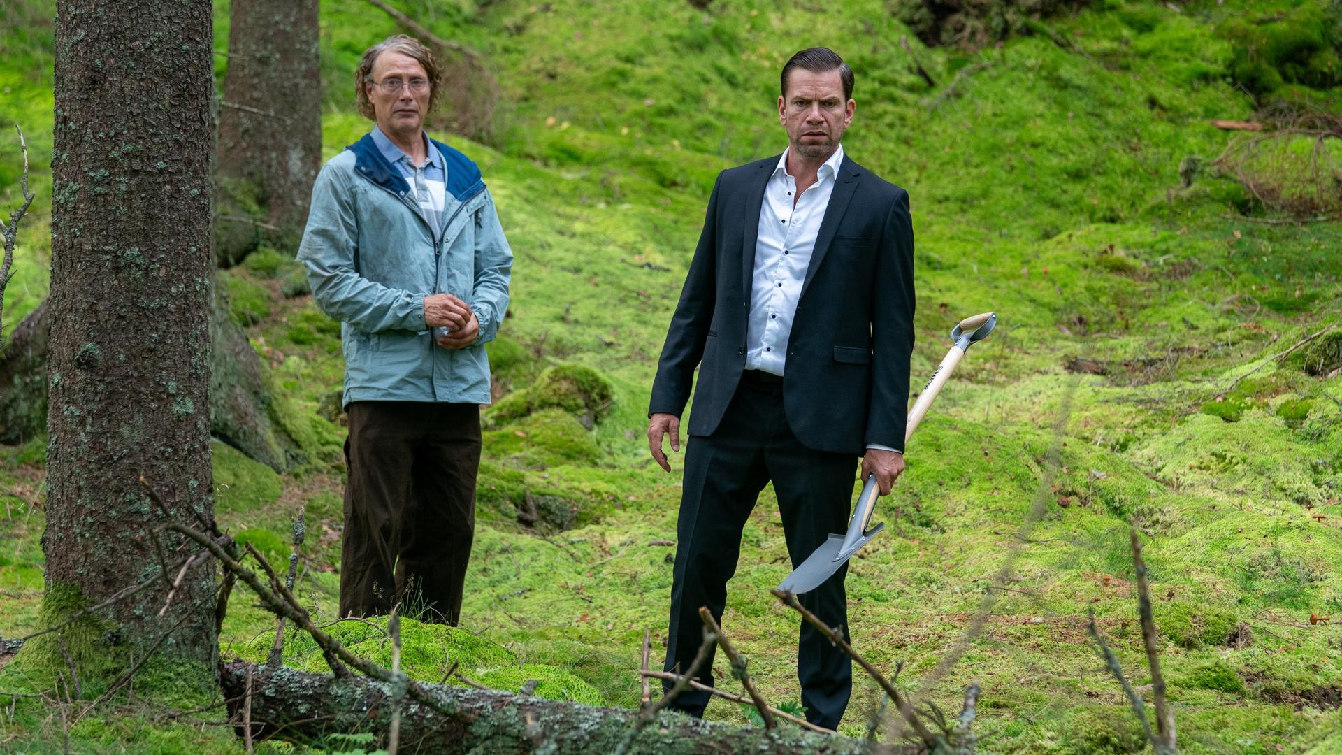 Zwei Männer stehen in einem moosbewachsenen Wald. Einer trägt einen schwarzen Anzug und hält eine Schaufel.