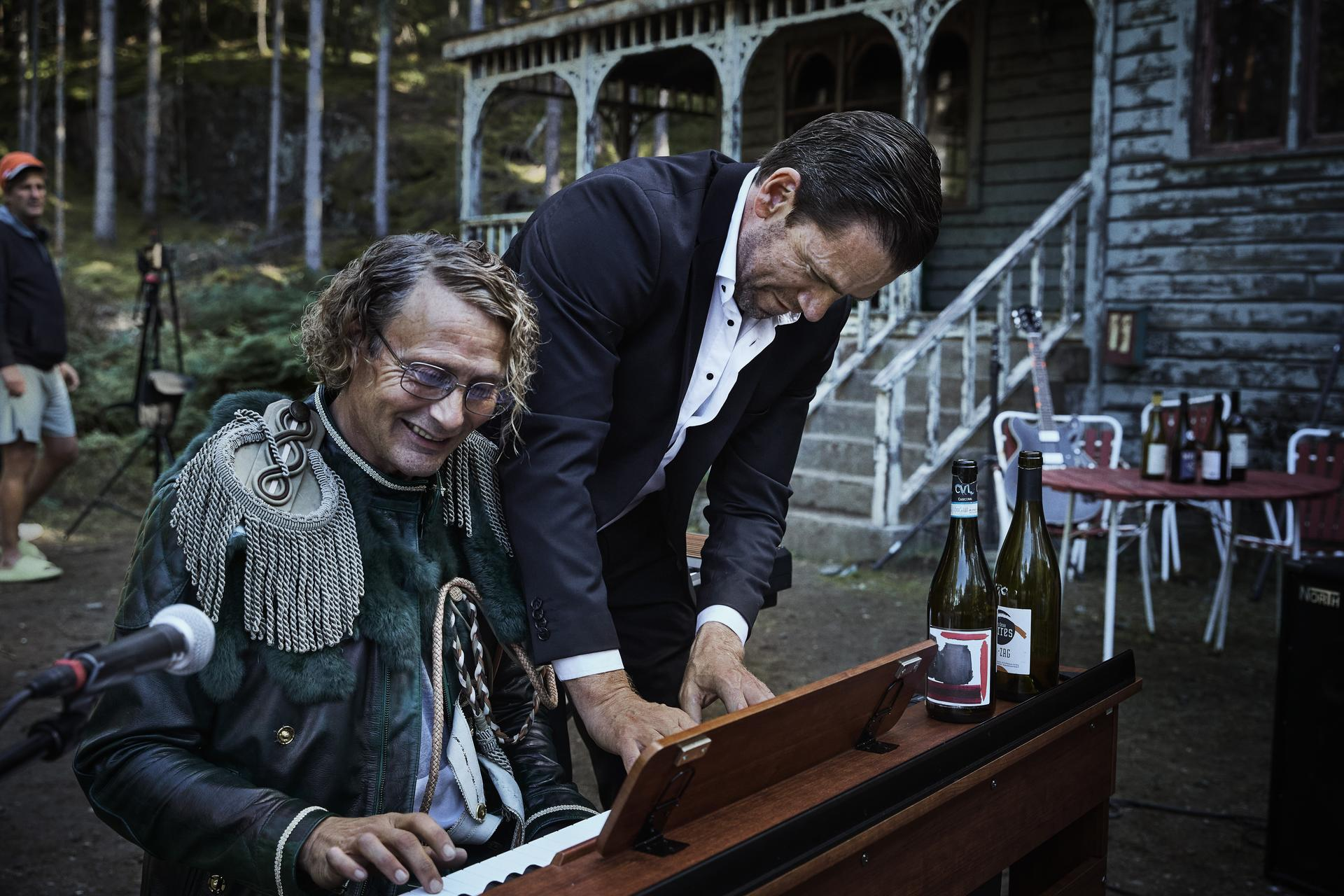 Ein Mann in Uniform spielt Klavier, ein anderer im Anzug steht daneben.