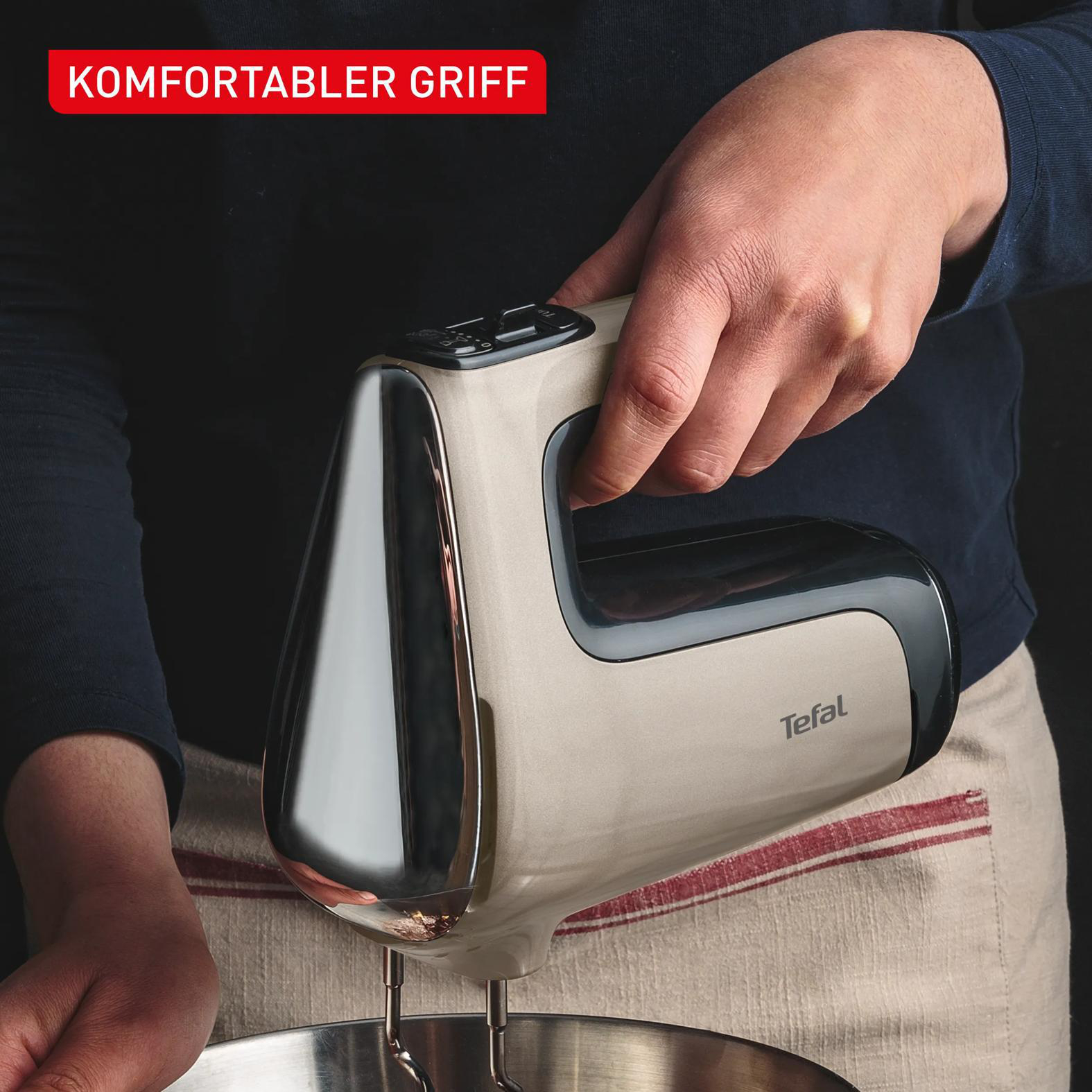Eine Hand hält einen beigefarbenen Handmixer mit polierten Metallaufsätzen über einer Schüssel.