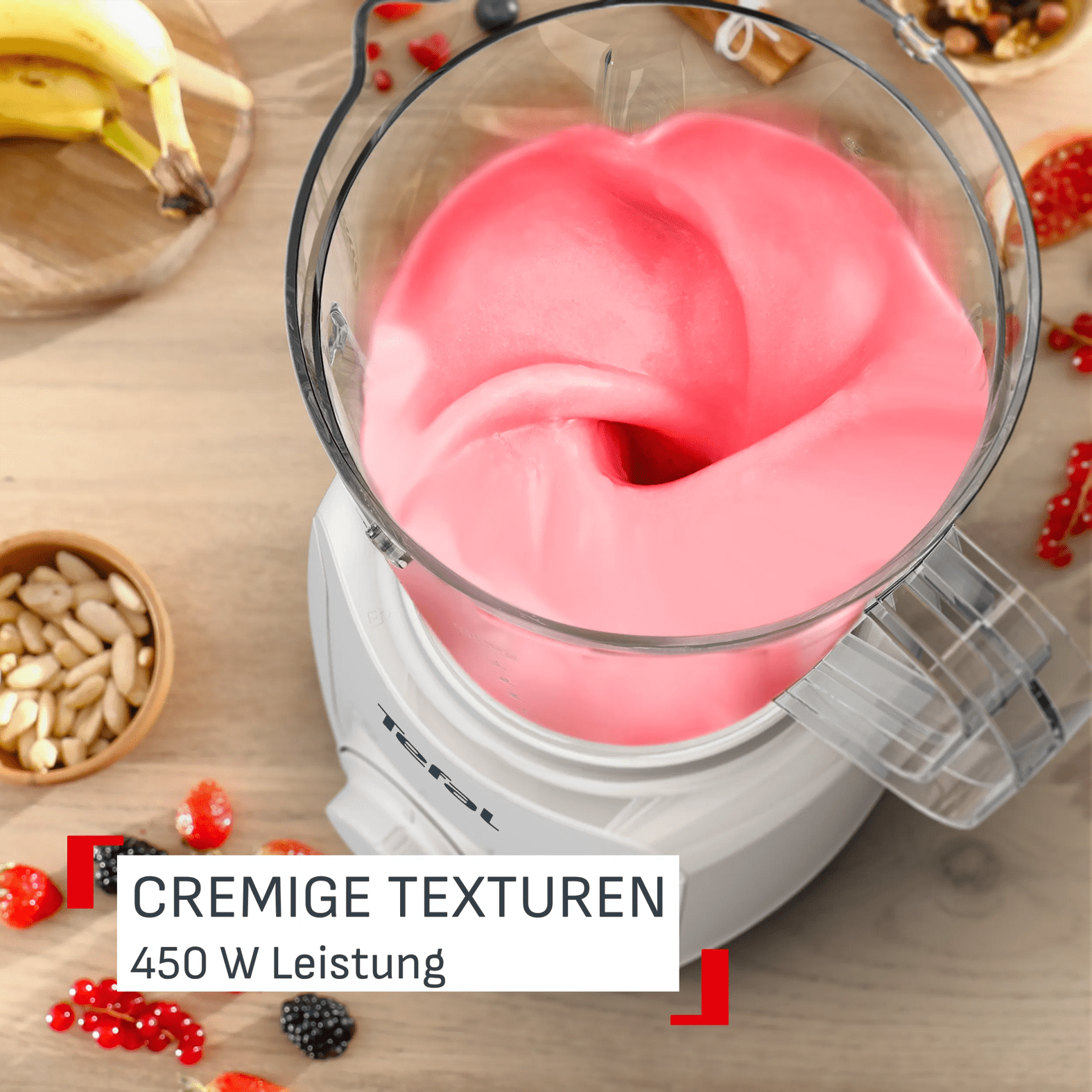 Ein Mixer mit cremiger rosa Masse, daneben Bananen, Beeren und Nüsse.
