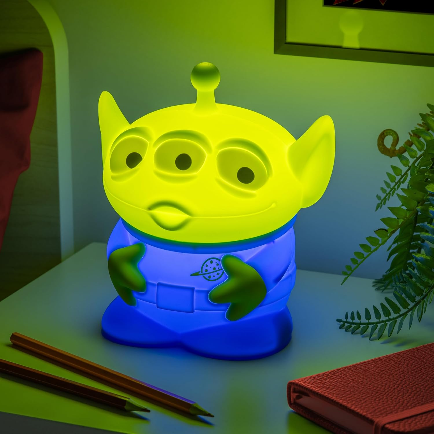 Sárga alien lámpa kéken világít egy éjjeliszekrényen, ceruzák és jegyzetfüzet mellett.