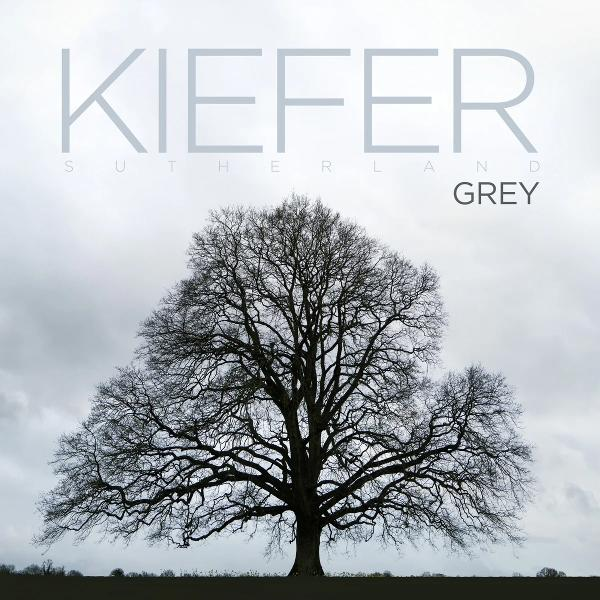 Ein großer kahler Baum steht vor einem bedeckten Himmel mit dem Namen KIEFER SUTHERLAND GREY.