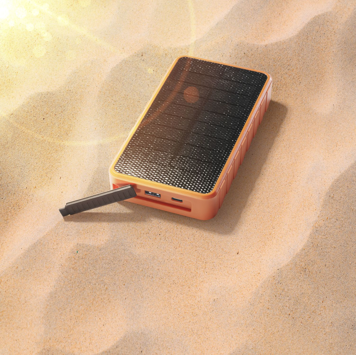 Batería externa naranja con panel solar y puertos USB sobre arena.