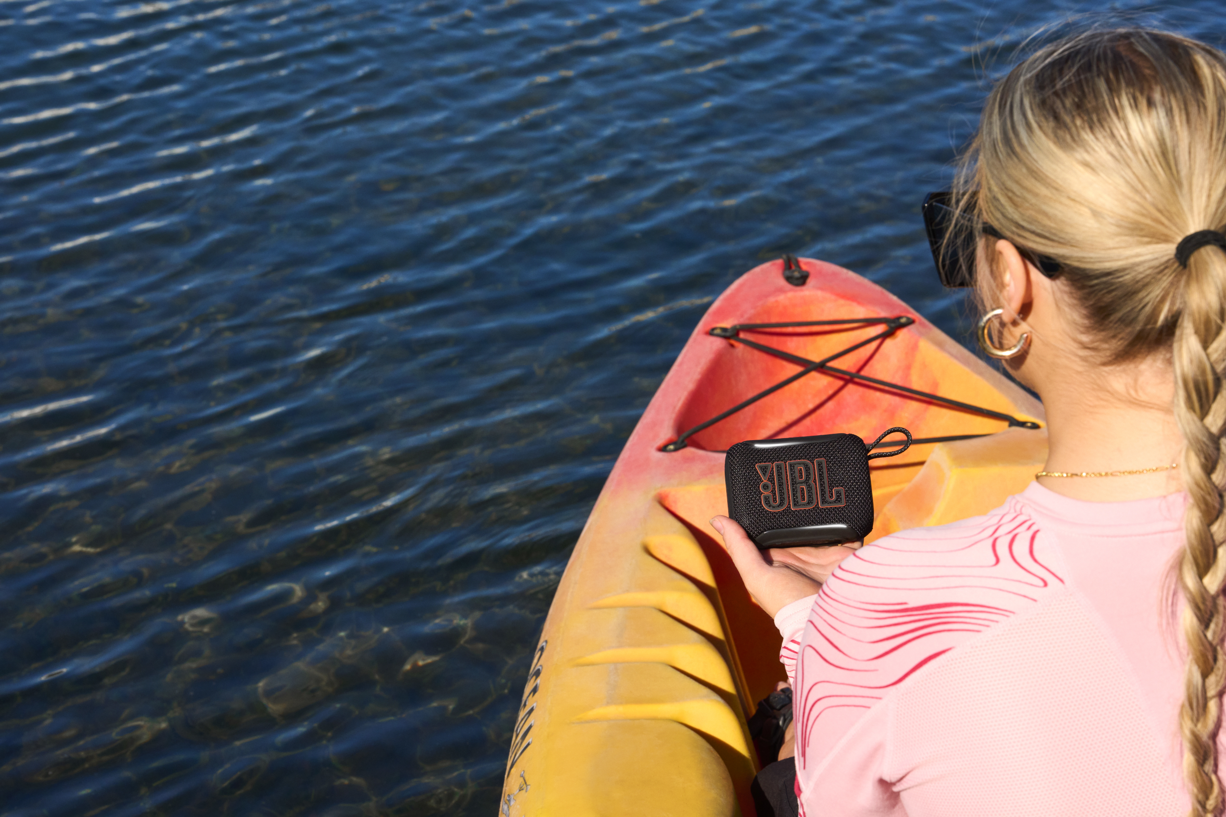 Frau auf einem Kajak hält eine schwarze JBL-Box über blauem Wasser.