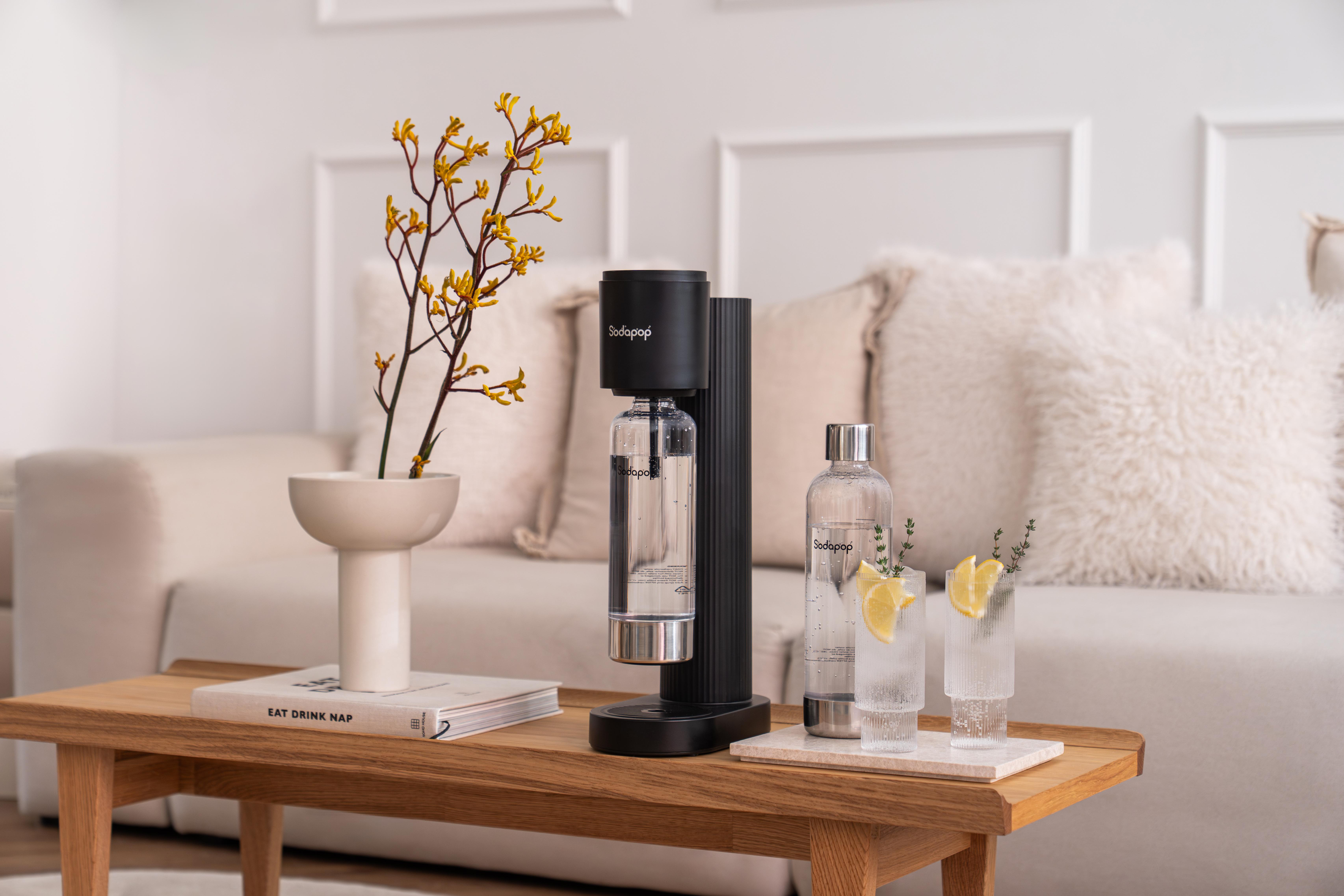 Ein schwarzer Sodastreamer steht auf einem Holztisch mit einem Buch und einer Vase mit gelben Blumen.