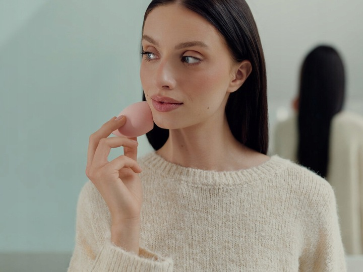 Kobieta z ciemnymi włosami trzyma różową gąbkę do makijażu blisko twarzy.