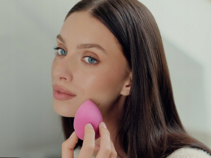 Kobieta z niebieskimi oczami trzyma w dłoni różową gąbkę do makijażu.