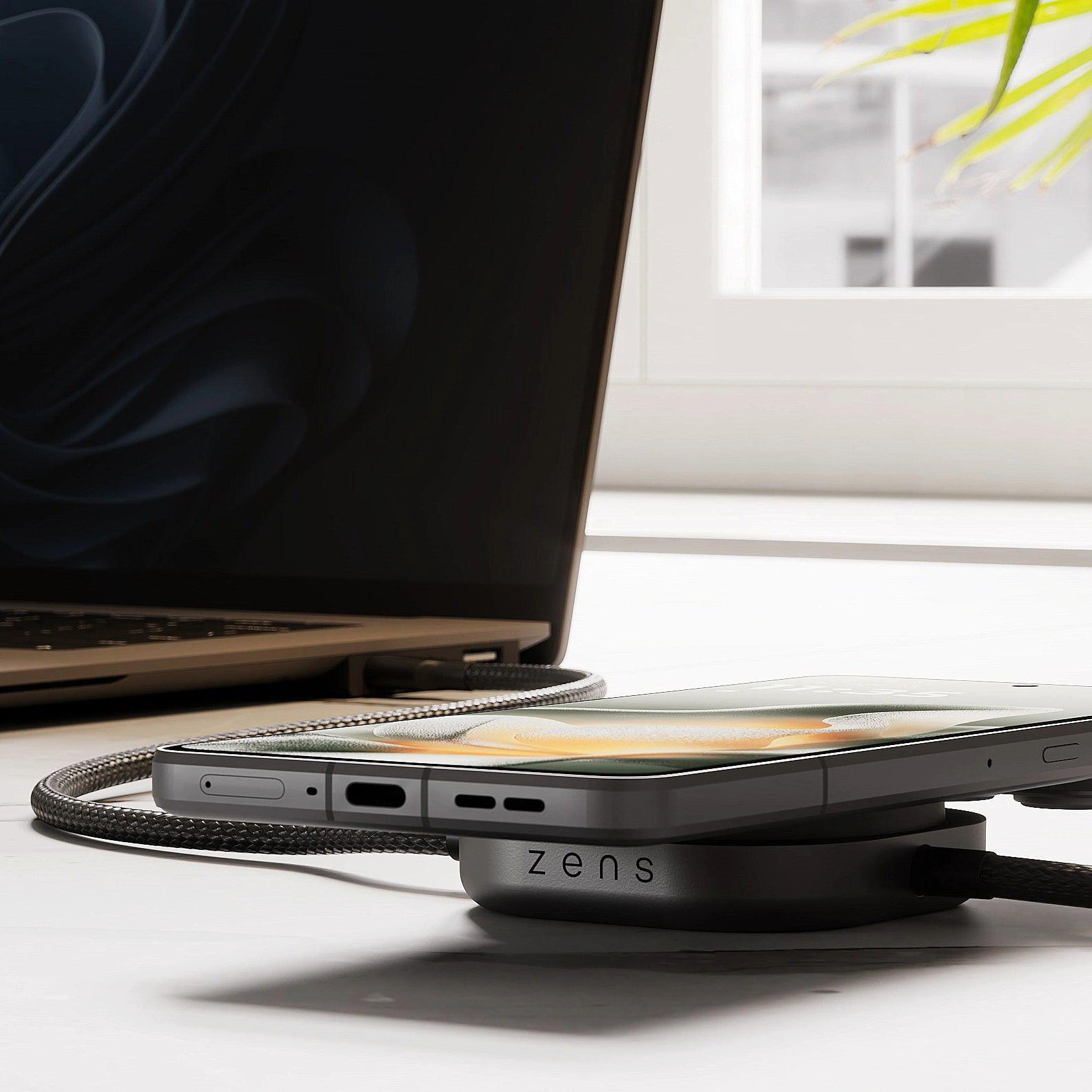 Ein Smartphone lädt kabellos auf einem Zens-Pad neben einem geöffneten Laptop.