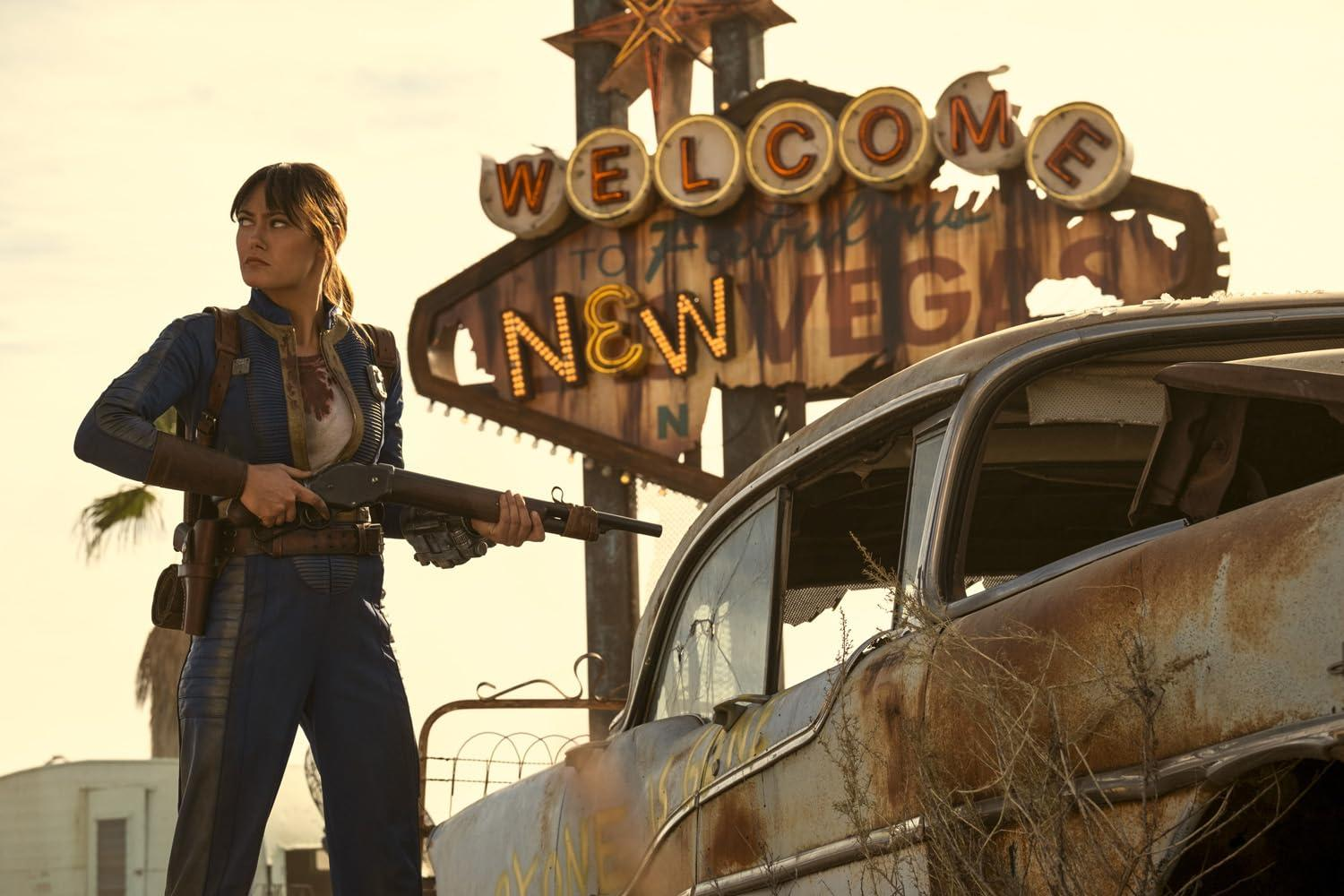 Frau mit Gewehr vor verrostetem Auto und "Welcome to New Vegas" Schild.