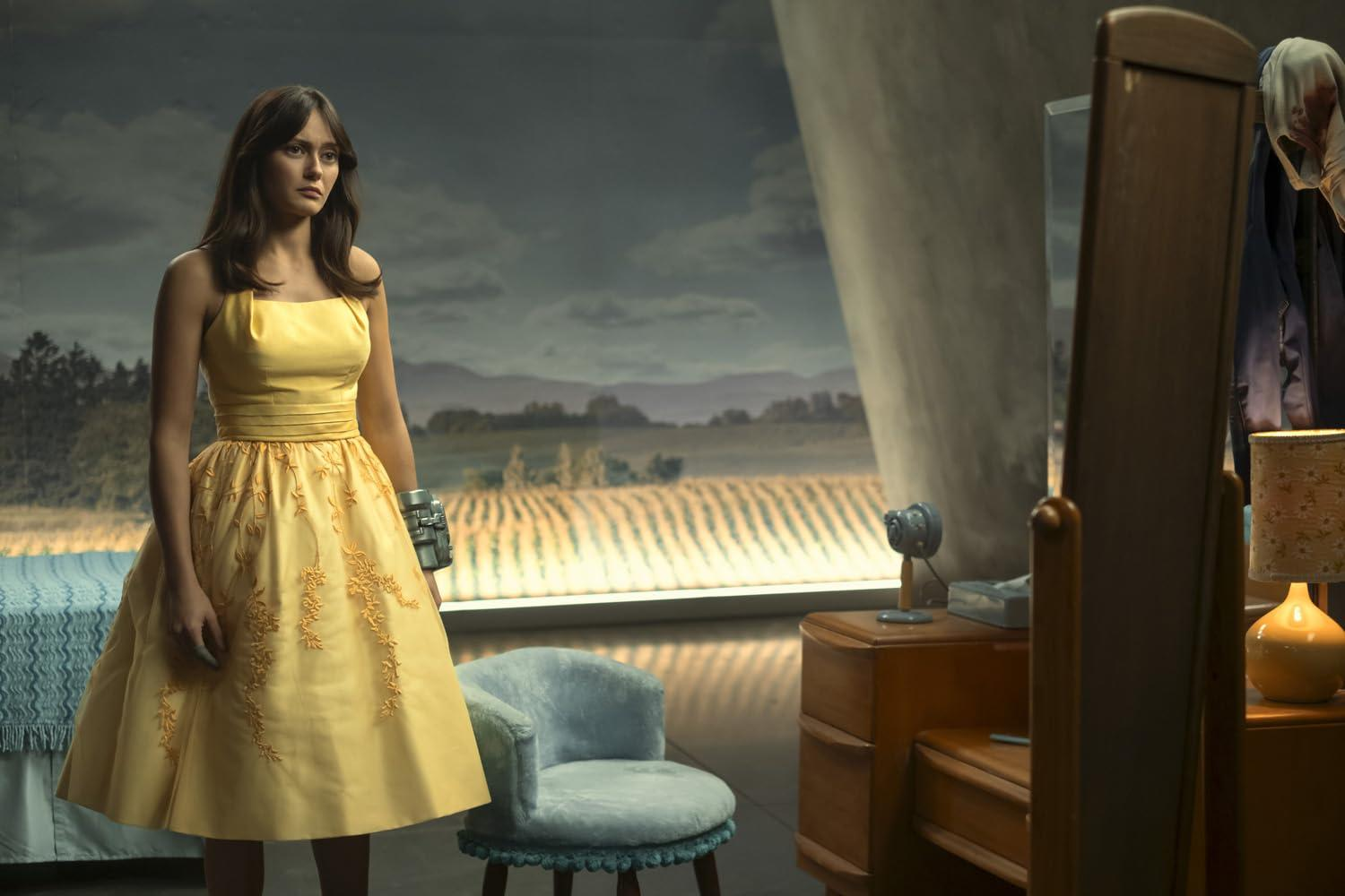 Frau in gelbem Kleid steht neben einem blauen Sessel vor einem Fenster.