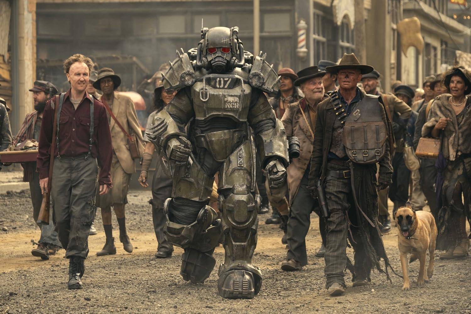 Eine Gruppe von Menschen und ein Hund gehen auf einer staubigen Straße, angeführt von einer Person in einem Power-Armor-Anzug.