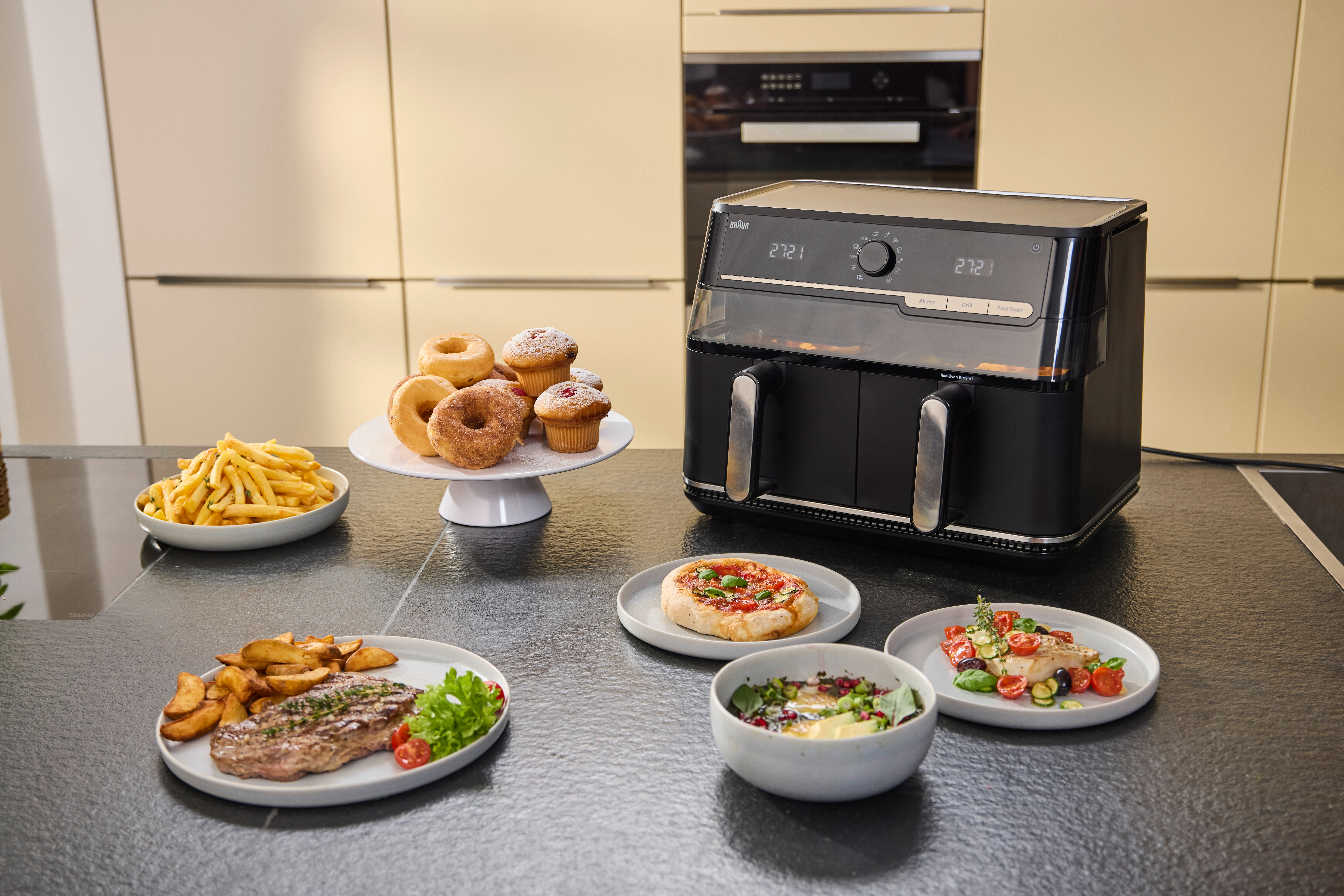 Ein schwarzer Heißluftfritteusen-Ofen neben Tellern mit Essen: Steak, Pommes, Pizza, Fisch und Gebäck.