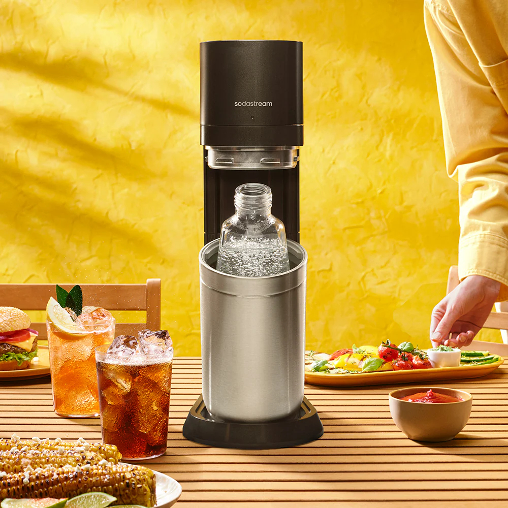 Una máquina Sodastream negra con una botella de agua está sobre una mesa con comida y bebidas.