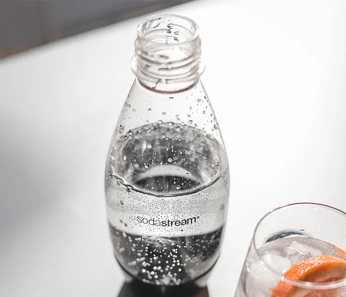 Una botella SodaStream con agua y burbujas junto a un vaso con hielo y pomelo.