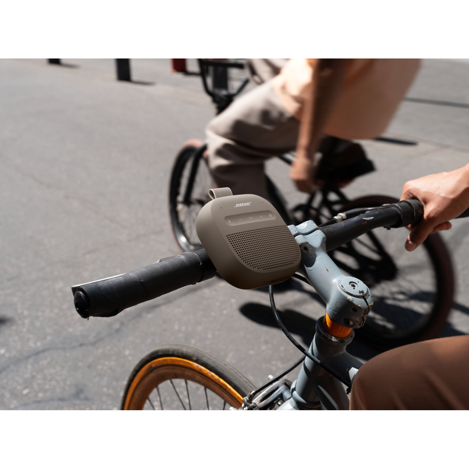 Een bruine Bose speaker is bevestigd aan het stuur van een fiets, met een andere fietser op de achtergrond.