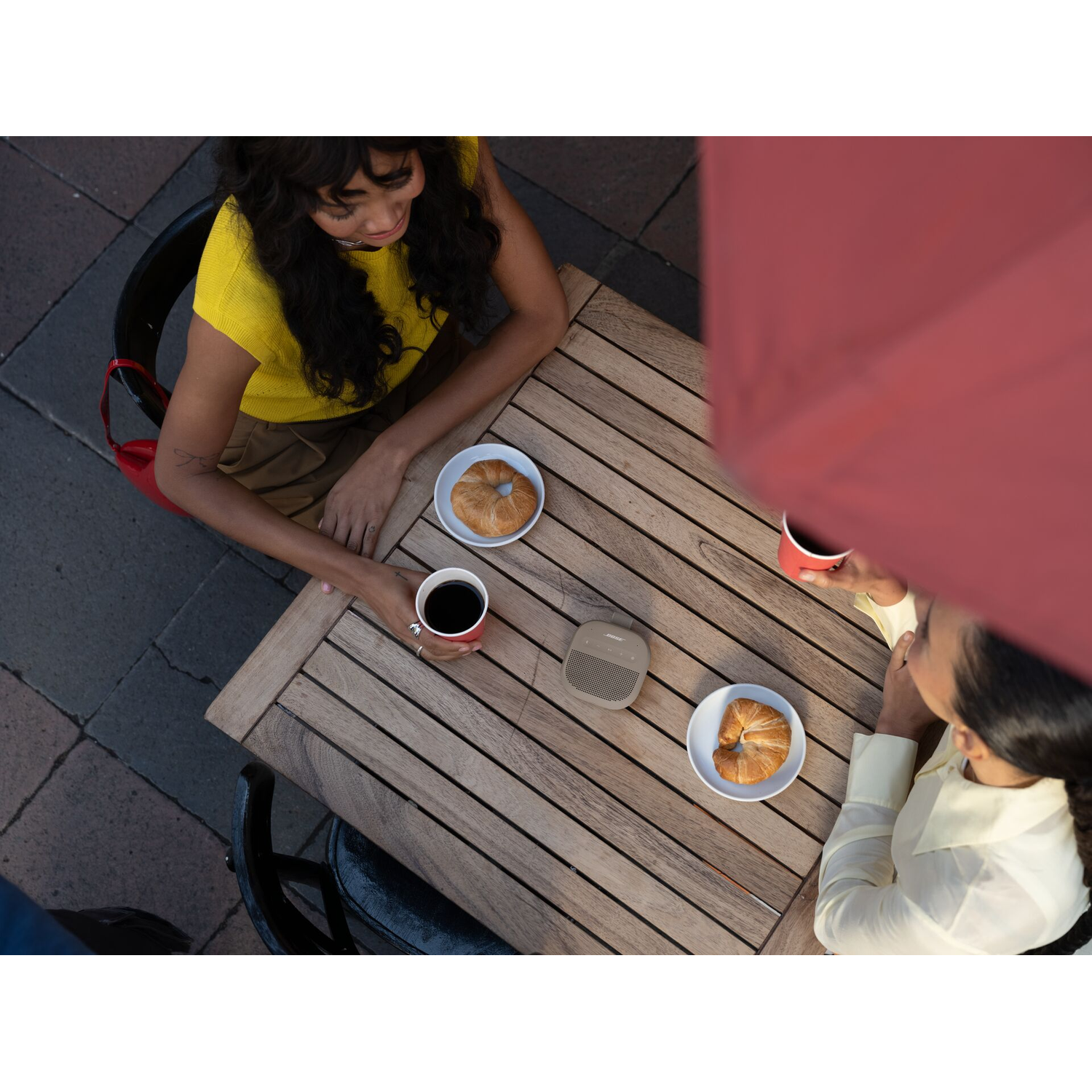 Twee vrouwen zitten aan een houten tafel buiten met een luidspreker en croissants.