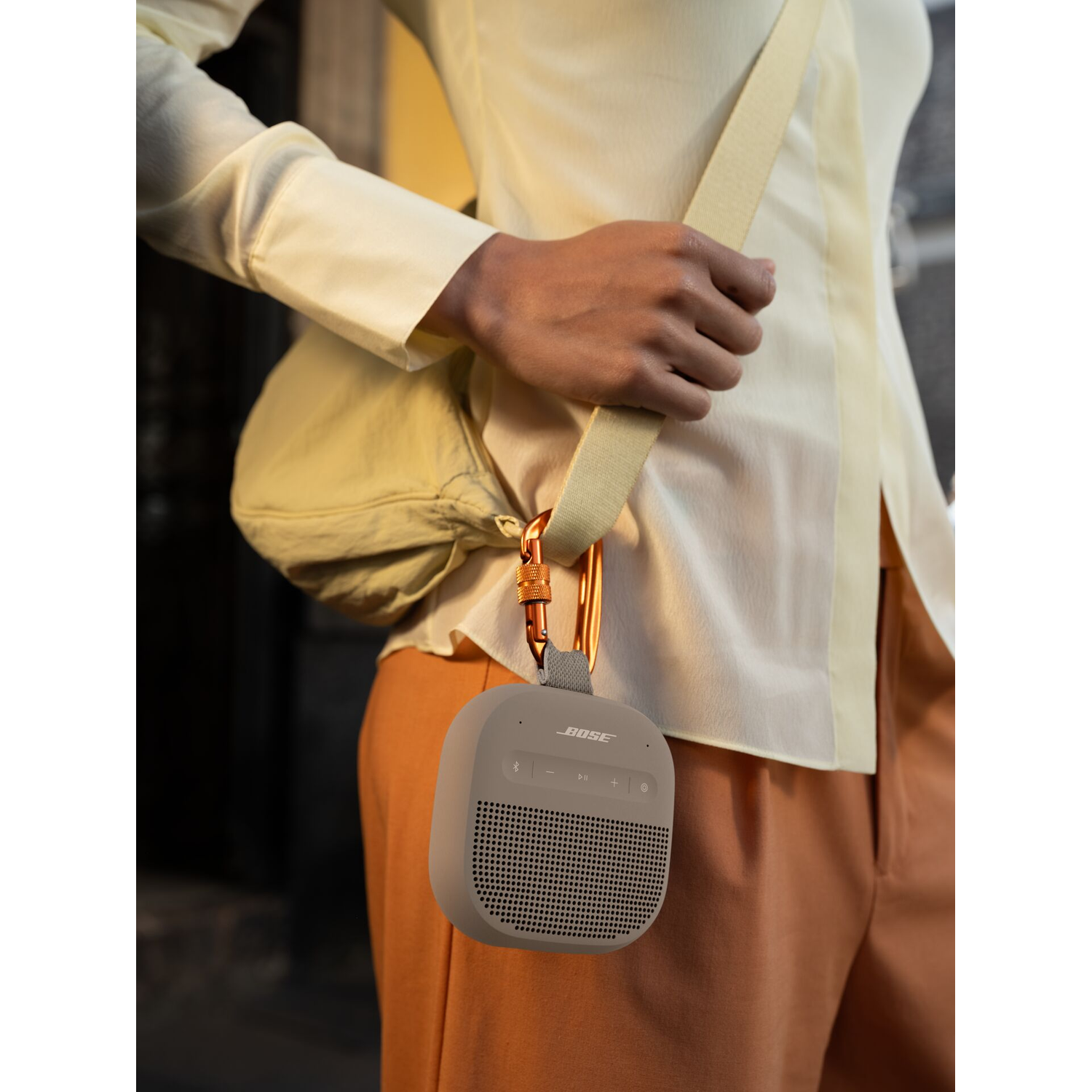 Grijze Bose speaker met riem en oranje karabijnhaak hangt aan een persoon in een lichtgekleurde blouse en broek.