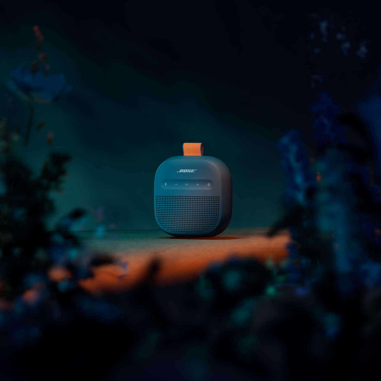 Donkerblauwe Bose speaker met oranje band op houten ondergrond, omringd door blauwe planten.