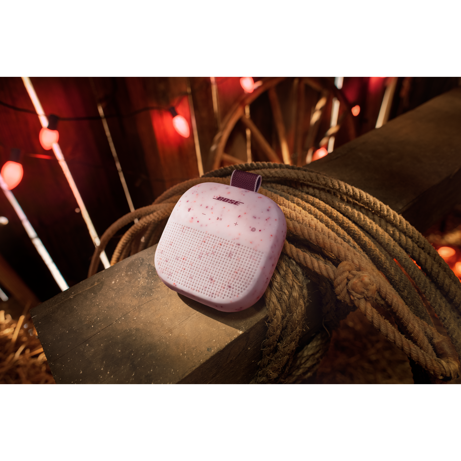 Een roze Bose speaker rust op een houten balk met touw en lampjes in een schuur.