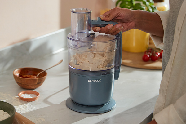 Eine Hand bedient eine Kenwood-Küchenmaschine mit Teig darin. Daneben sind Schüsseln.