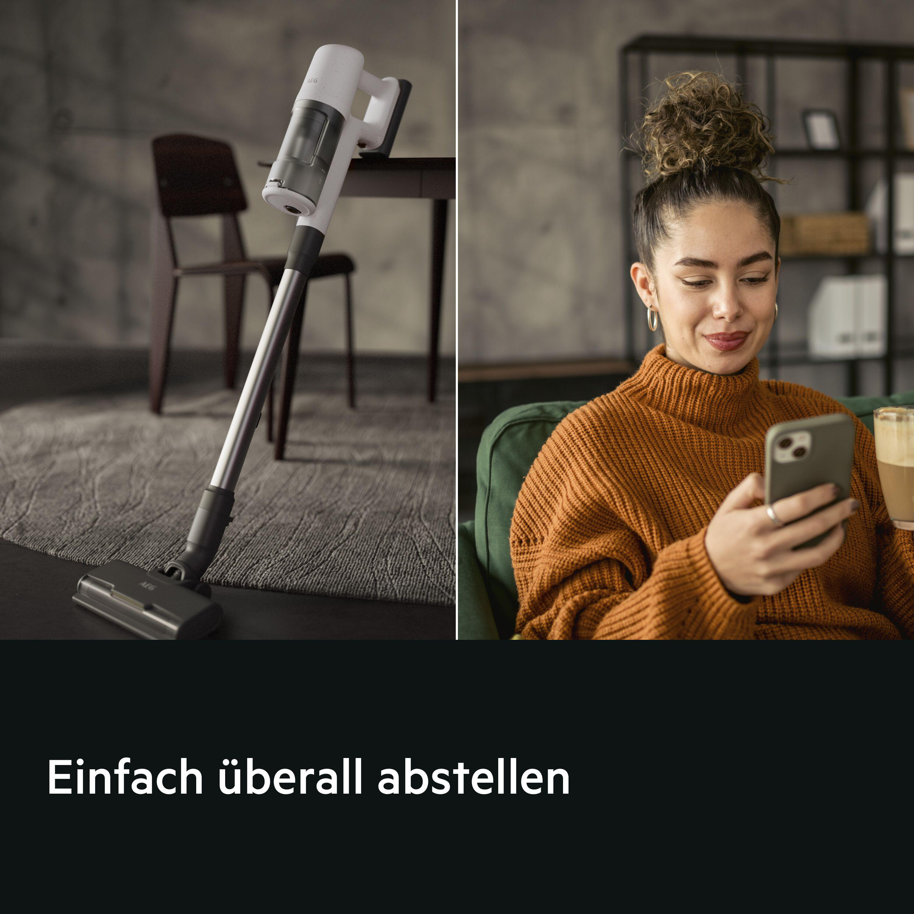 Ein weißer Handstaubsauger steht auf einem Teppich, eine Frau benutzt ihr Handy.