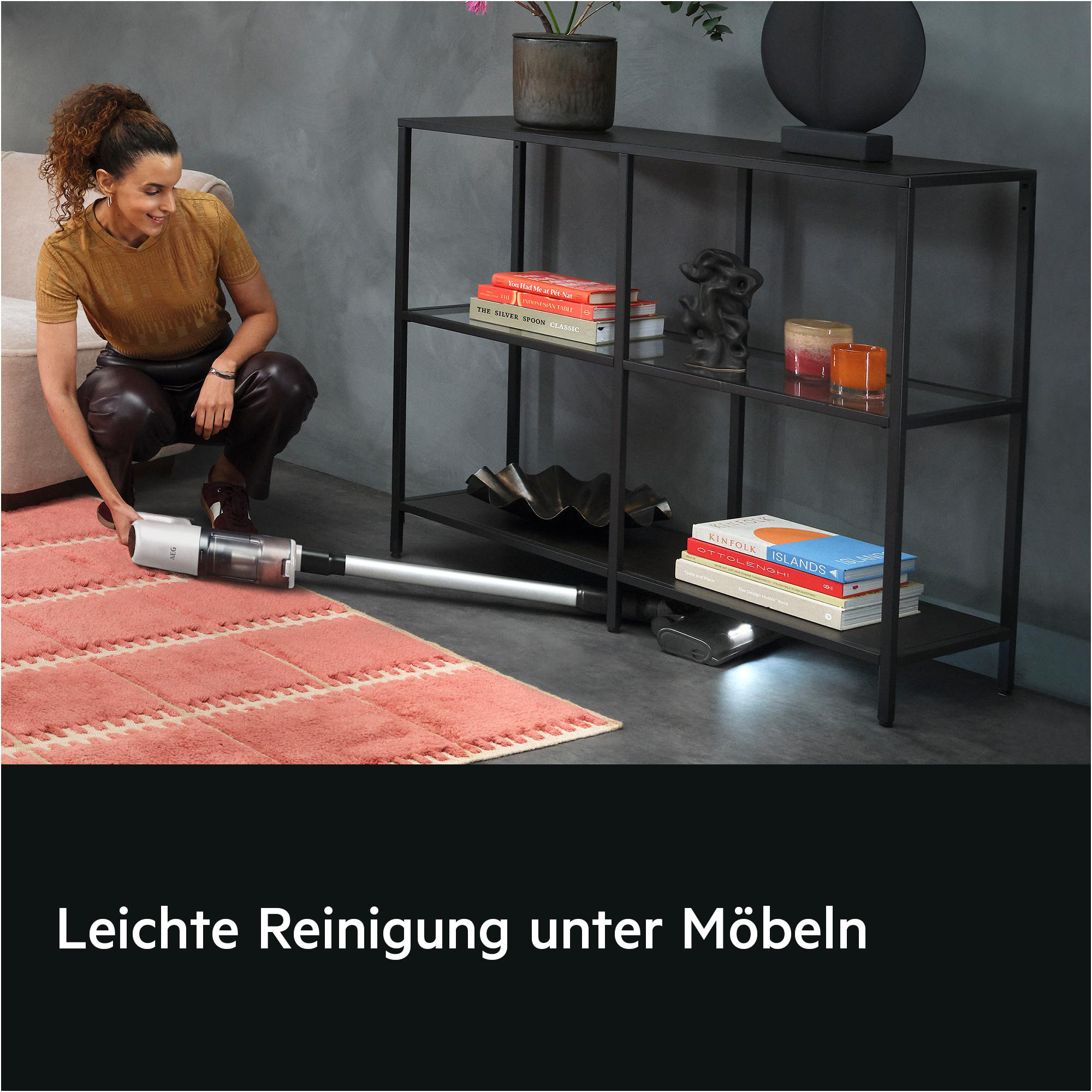 Eine Frau reinigt Teppich unter Möbeln mit einem Stabstaubsauger.