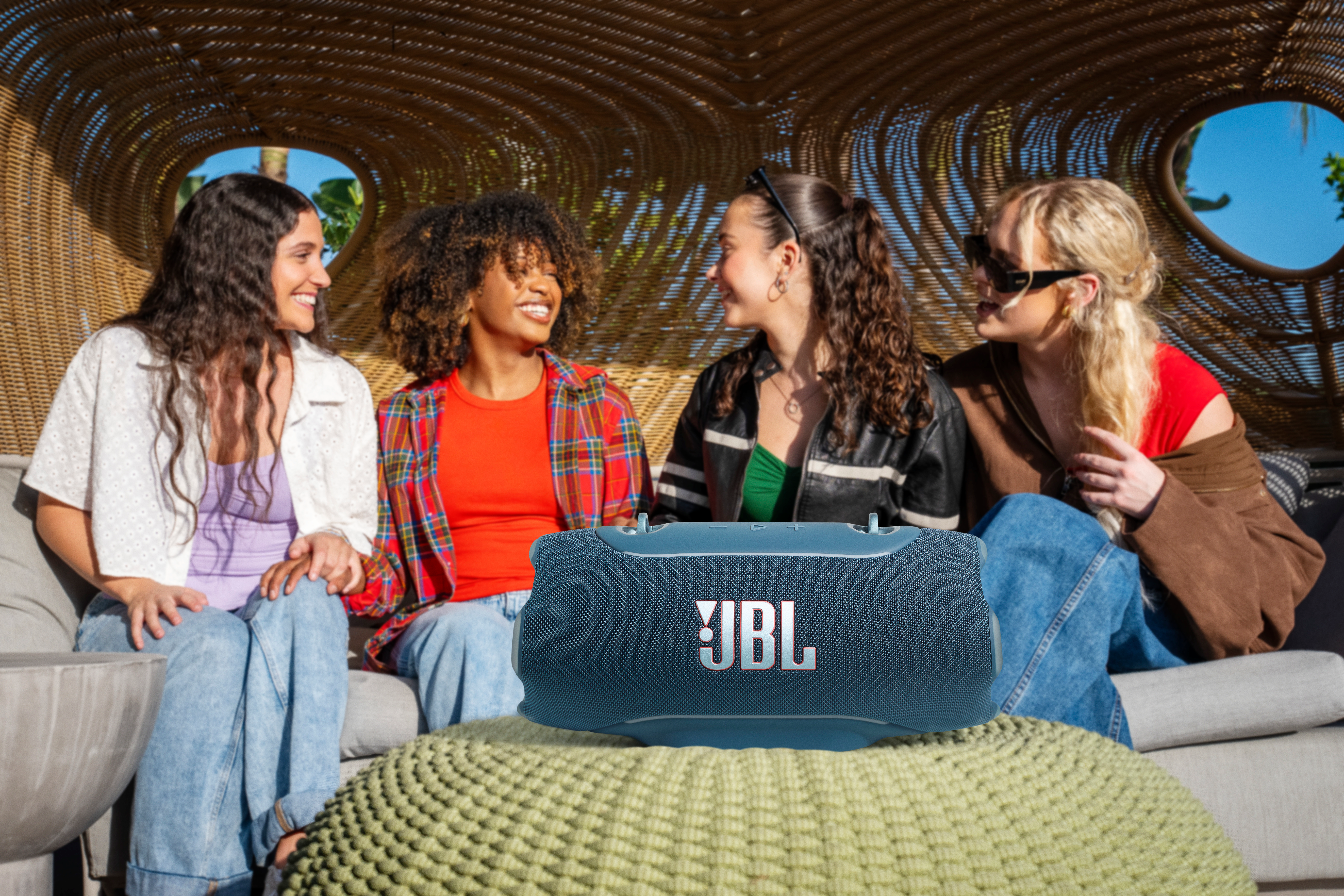 Vier junge Frauen sitzen auf einer Couch und lachen. Eine blaue JBL-Box steht davor.