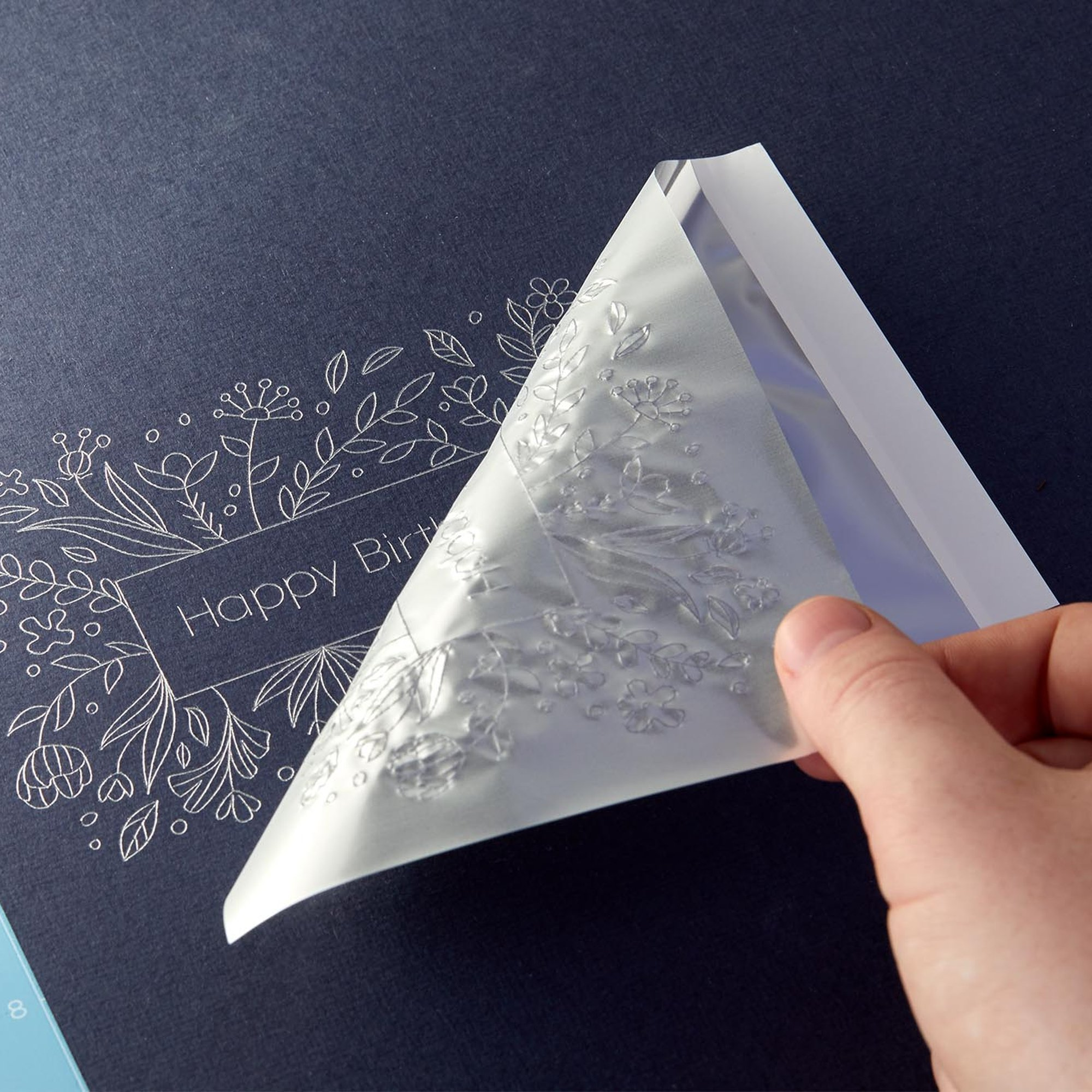 Mano retira película protectora de tarjeta con diseño floral y texto 'Feliz Cumpleaños'.