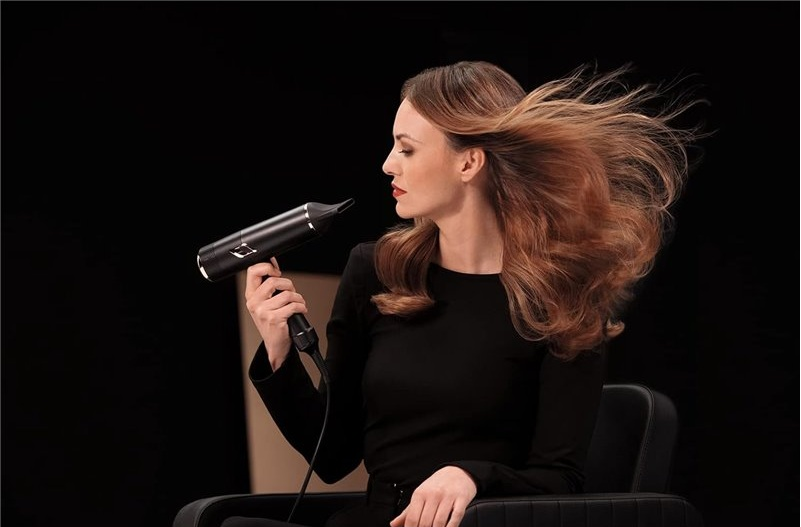 Kobieta z brązowymi włosami suszy włosy czarną suszarką. Nosi czarną sukienkę.