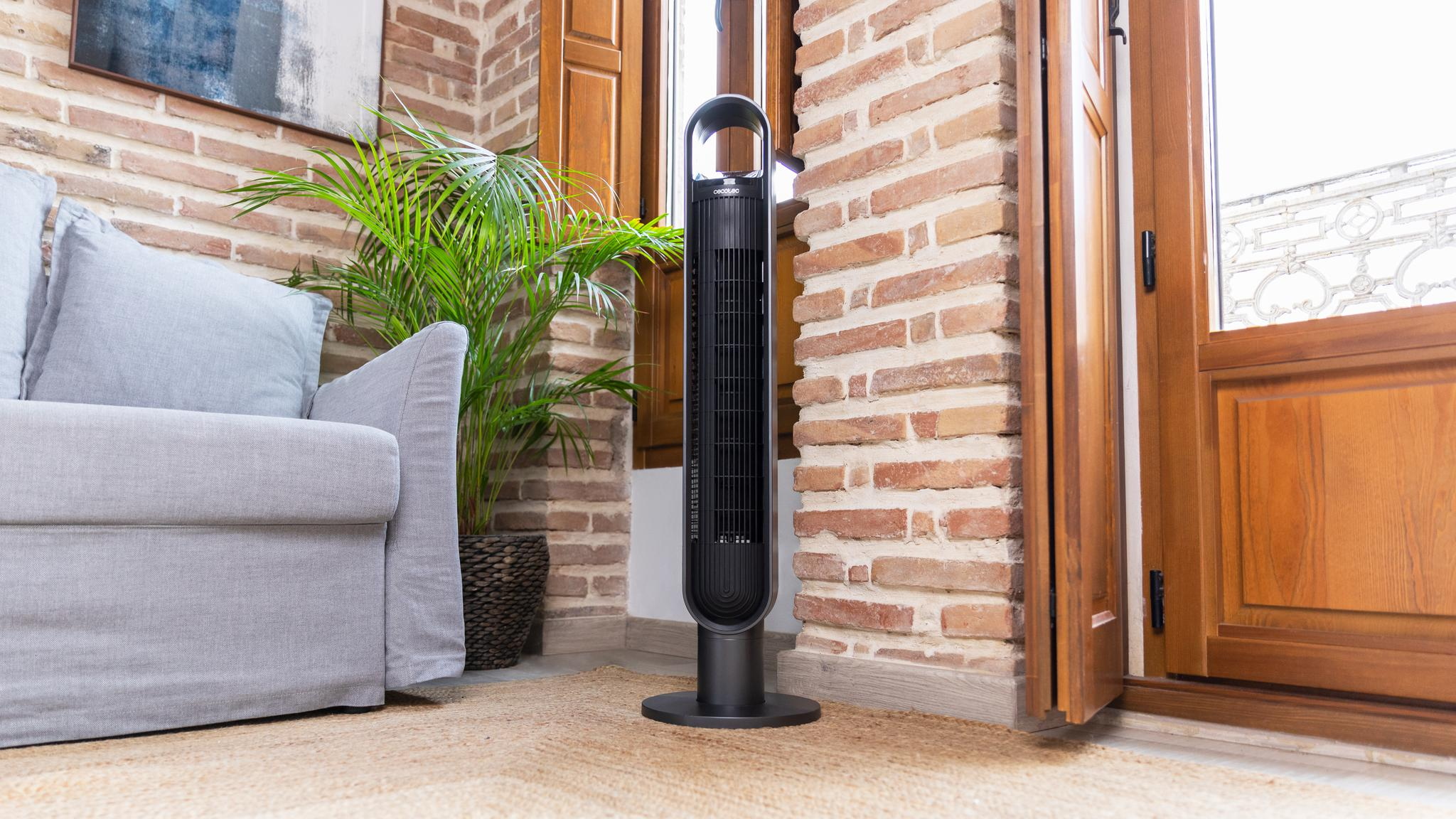 Un ventilador de torre negro y delgado junto a un sofá gris y una palmera.