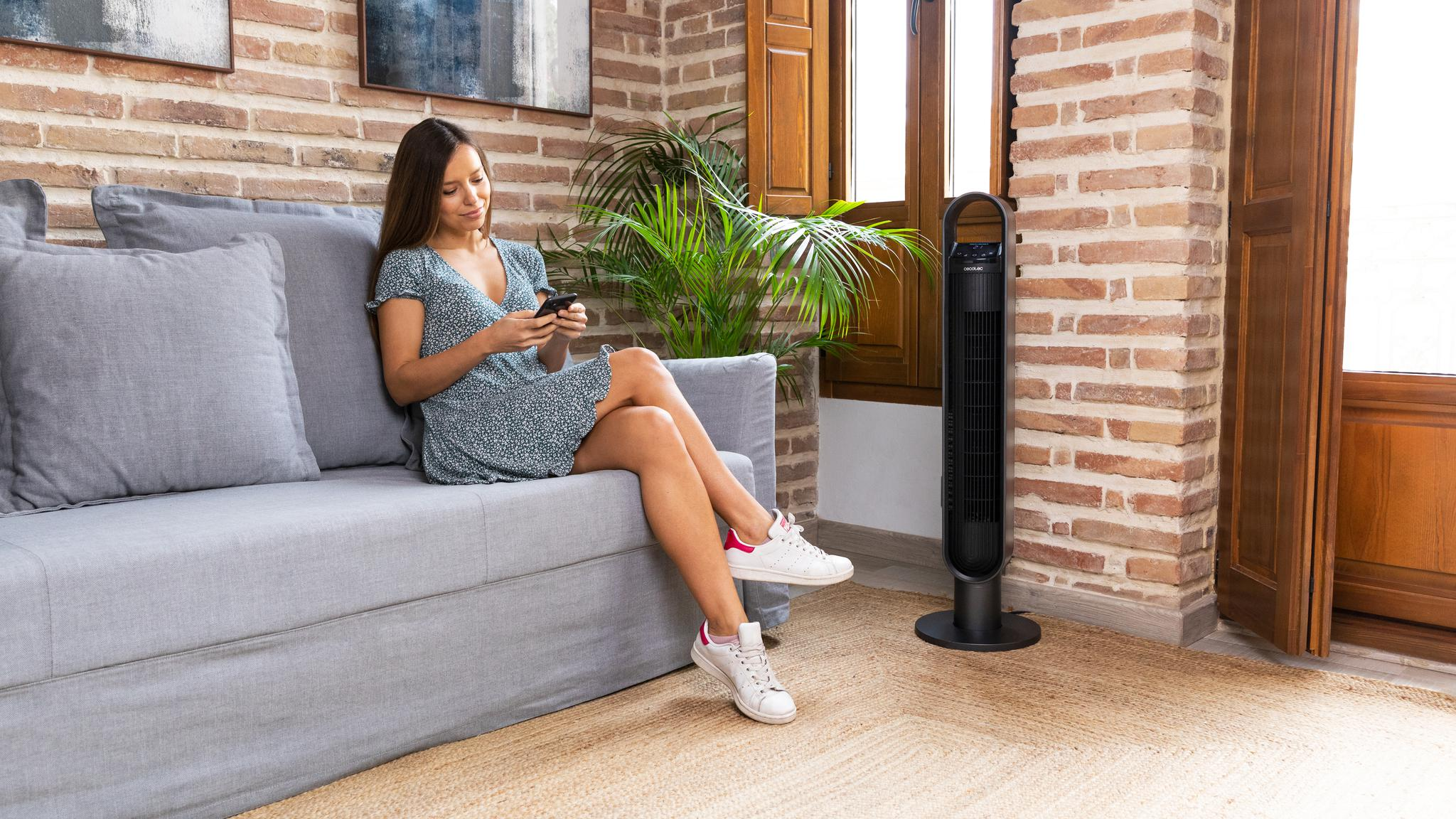 Una mujer sentada en un sofá mira su teléfono, un ventilador de torre a su lado.