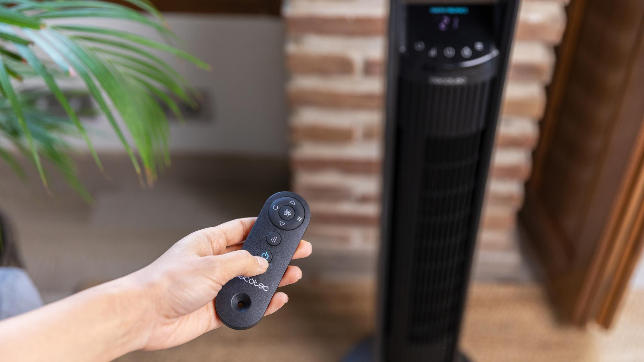 Mano sujetando mando negro, pulsando botón de encendido. Ventilador torre negro al fondo.