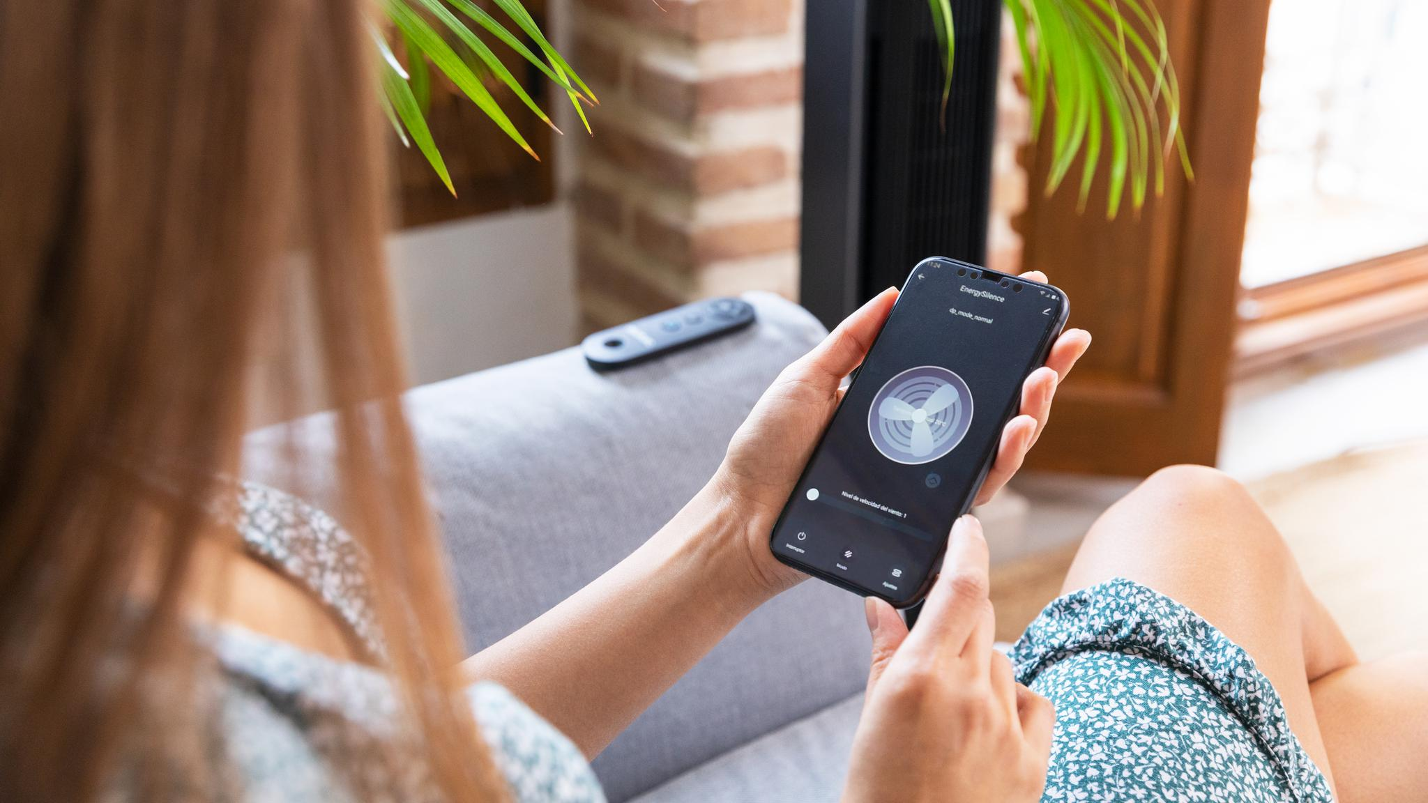 Mujer sostiene teléfono con app de ventilador; control remoto en sofá.