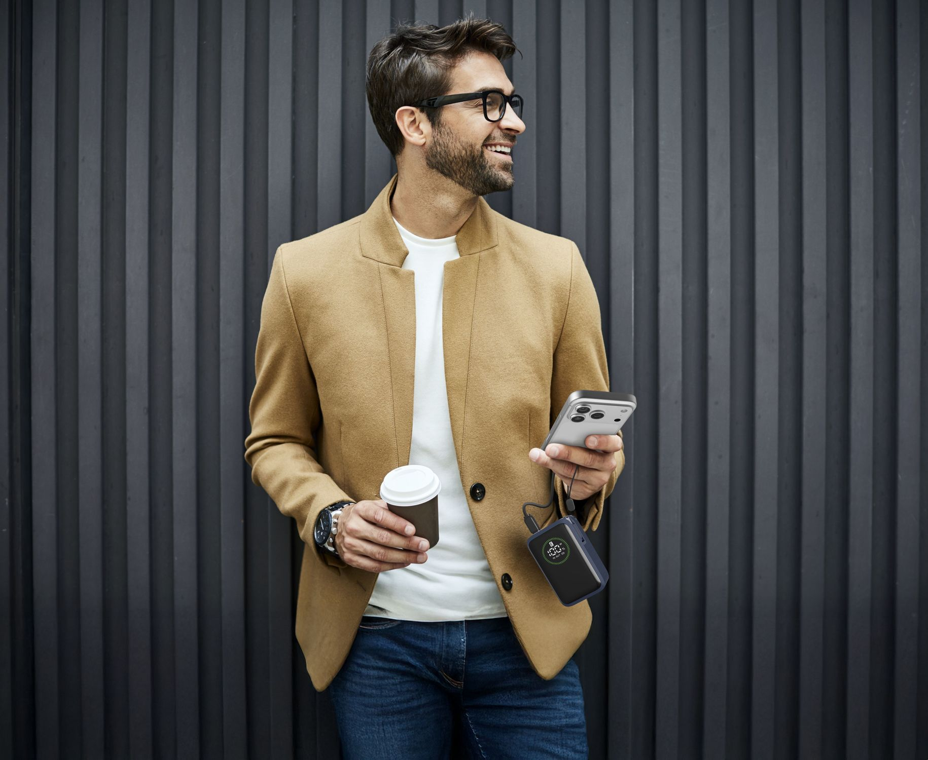 Un hombre con gafas sostiene café y un móvil con cargador contra pared oscura.