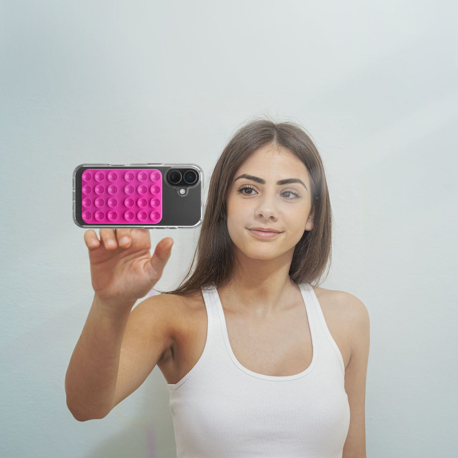 Una mujer con camiseta blanca sostiene un smartphone con funda pop-it rosa sobre fondo azul claro.