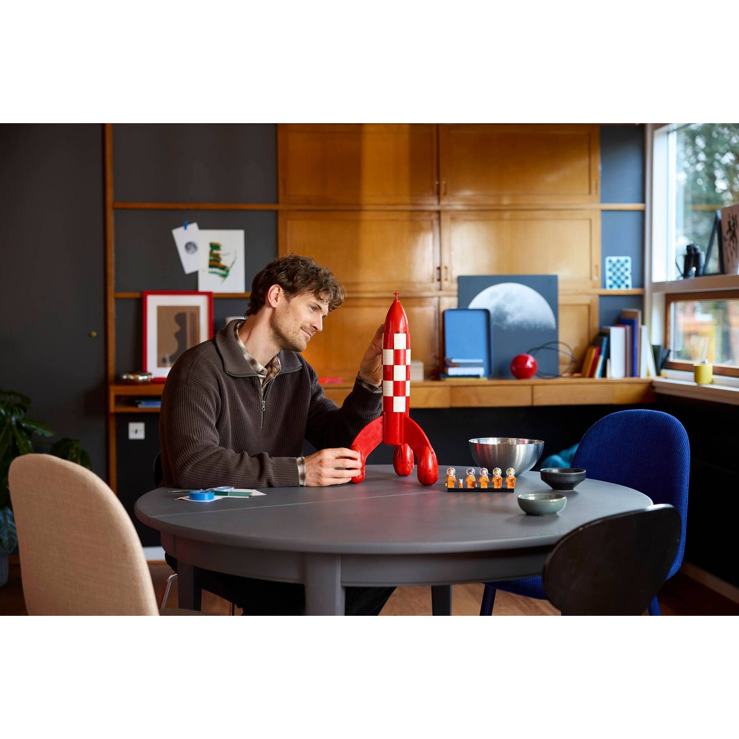 Man assembling a red rocket model kit with figures on a round table.