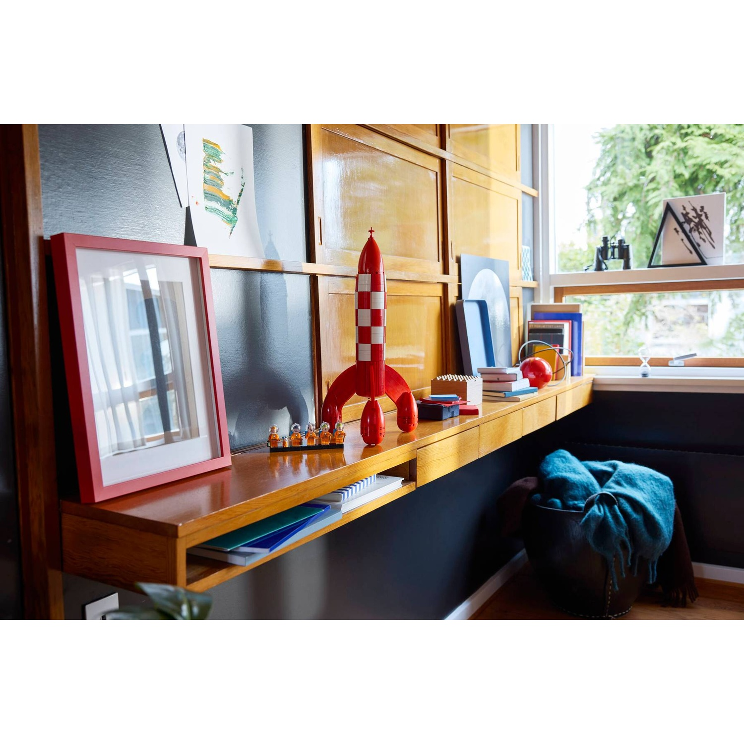 A red rocket toy sits on a wooden desk with books and framed art.