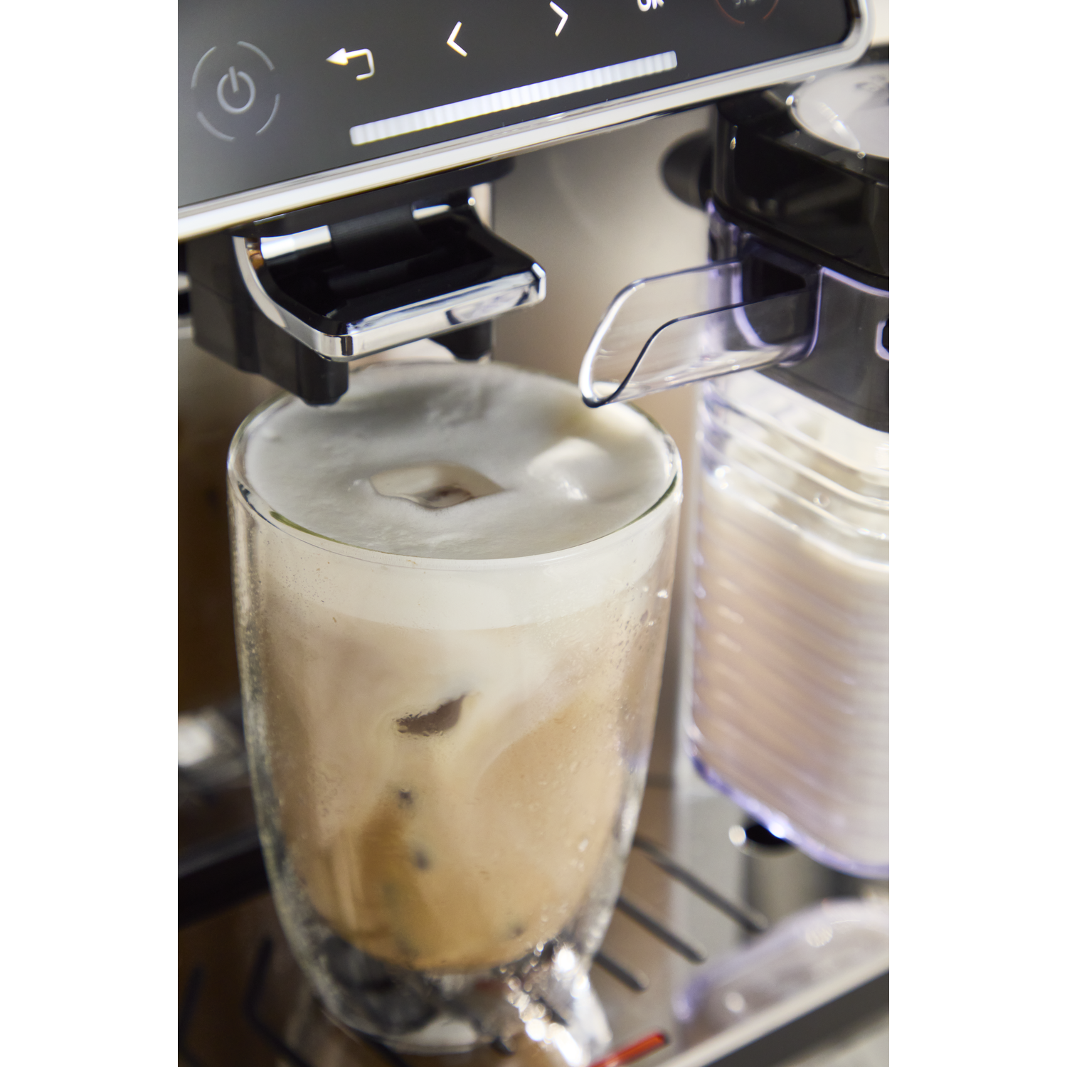 Close-up van een glas ijskoffie met schuim en ijs naast een koffiezetapparaat.