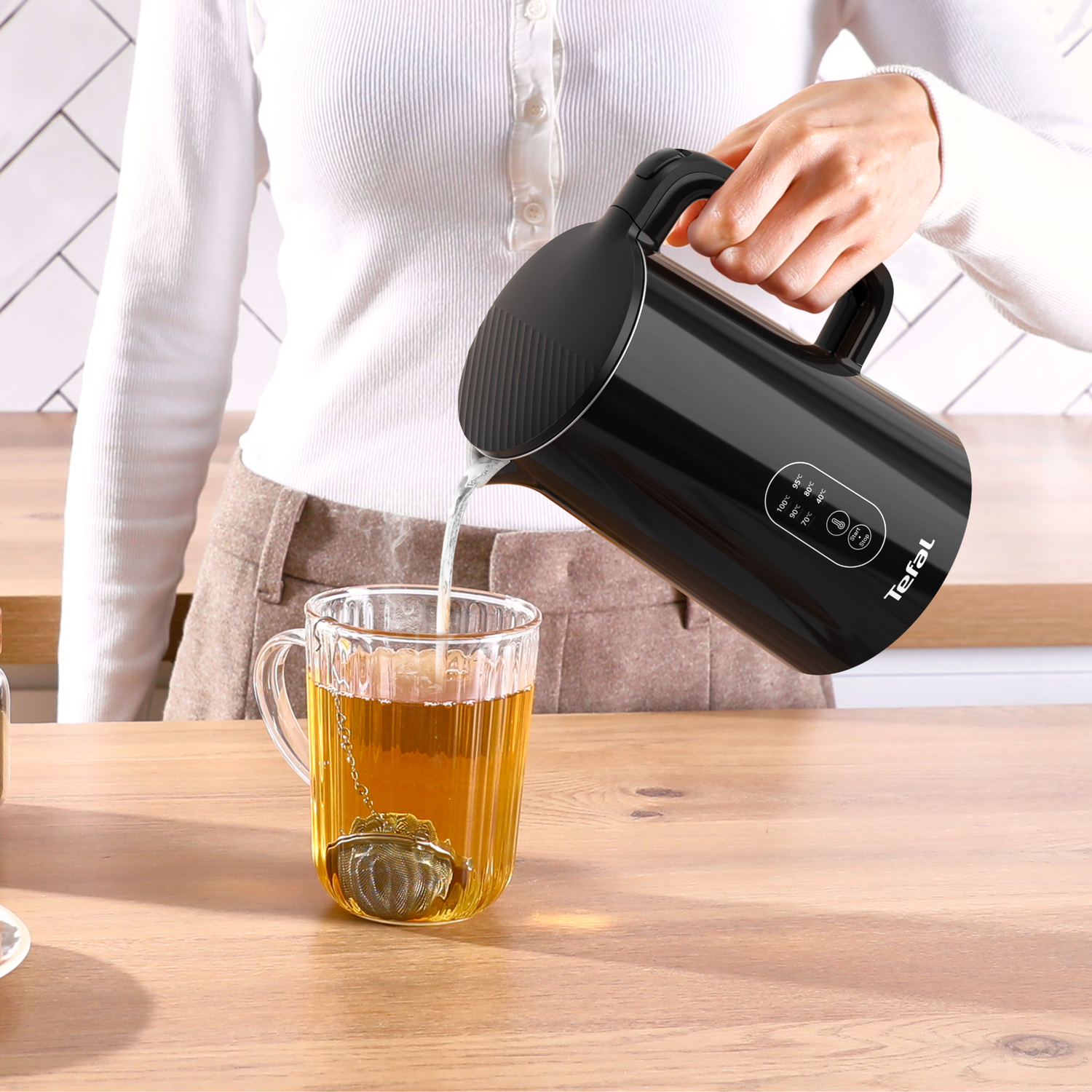 Een persoon schenkt heet water uit een zwarte Tefal waterkoker in een glazen mok met thee-infuser.