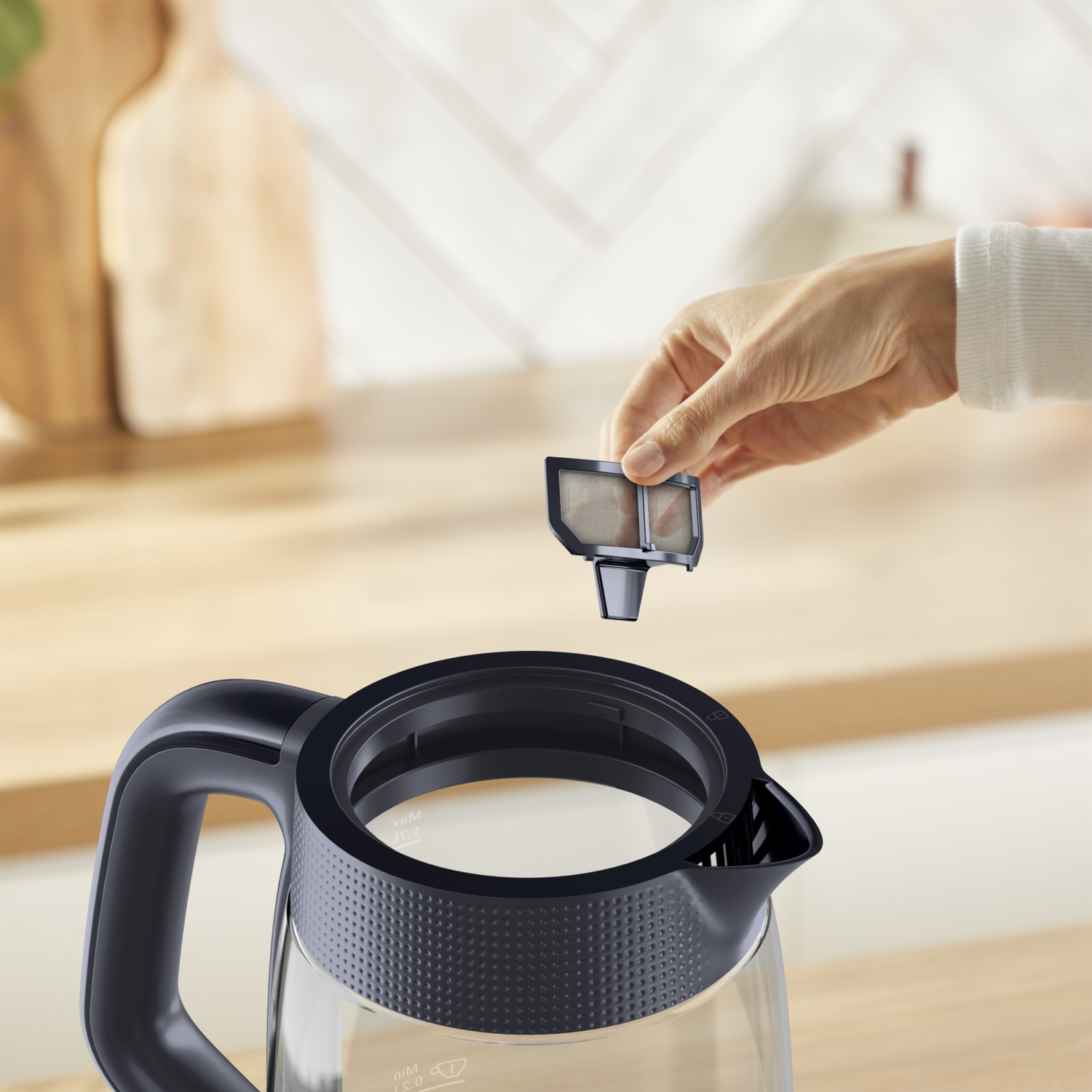 Een hand verwijdert een filter uit een glazen waterkoker met zwarte handgreep.