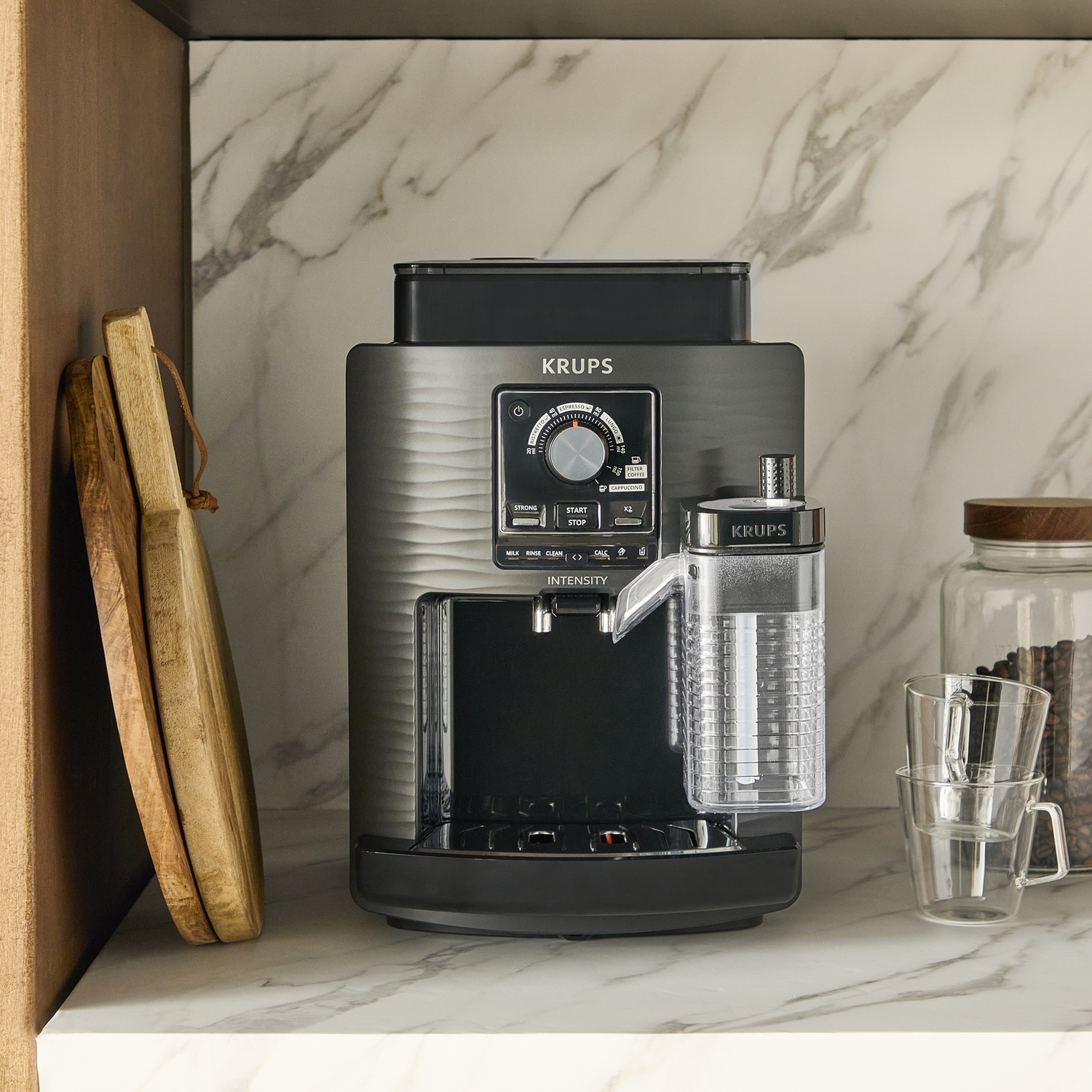 A Krups espresso machine with milk container, cups, and wooden cutting boards.