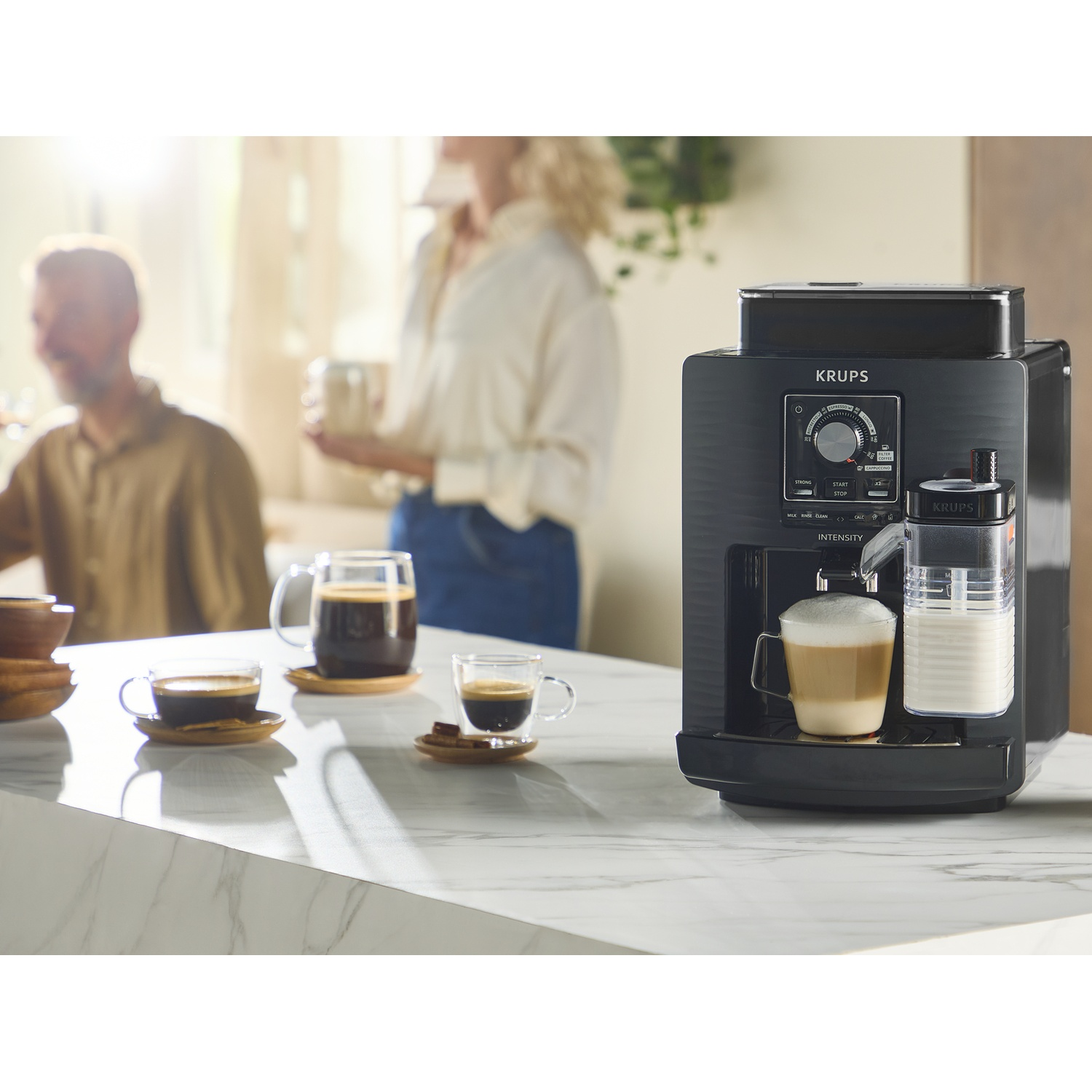 Coffee machine brews cappuccino, two people blurred in background.