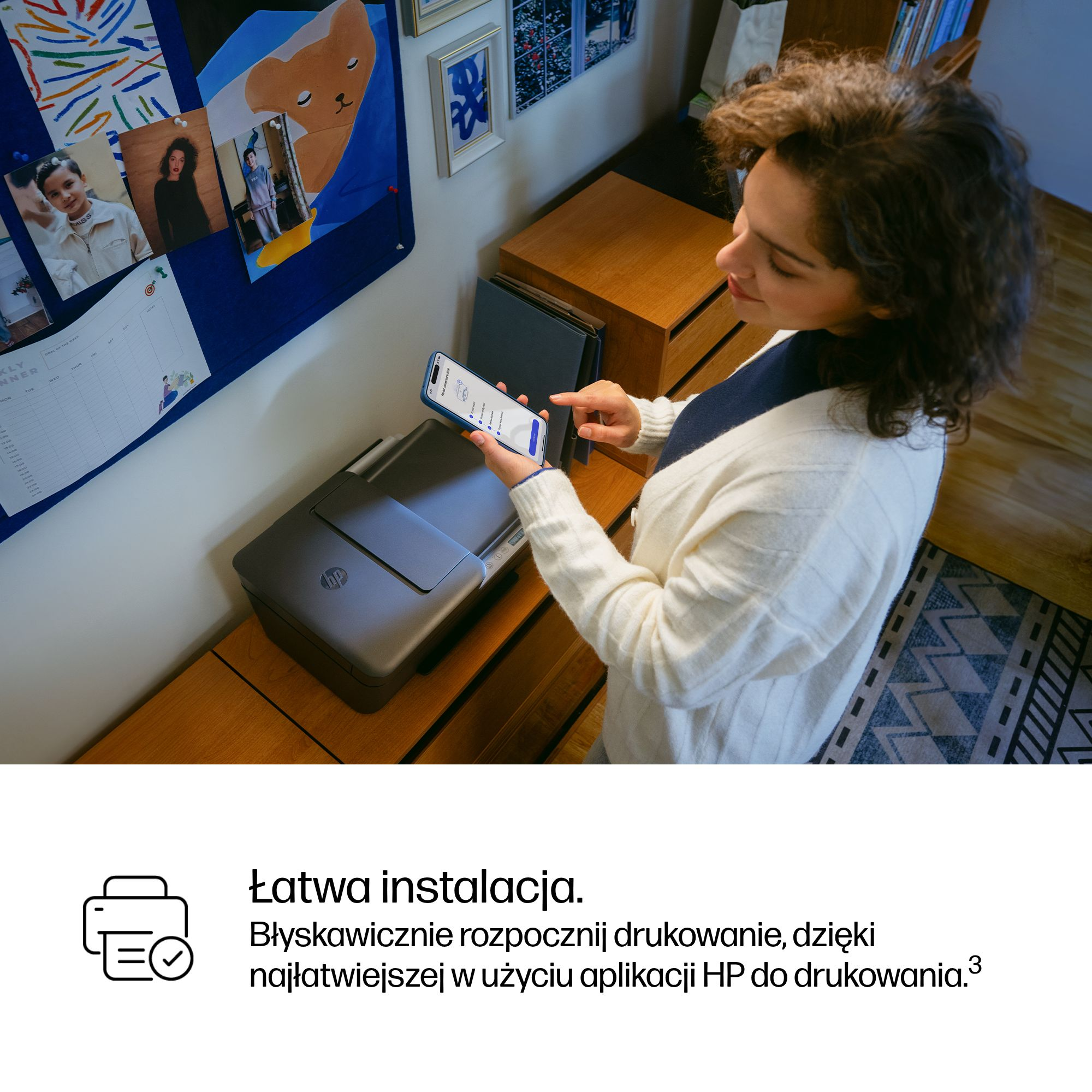 Kobieta trzyma smartfon, kierując go na drukarkę. Po lewej stronie znajduje się tablica ogłoszeń.