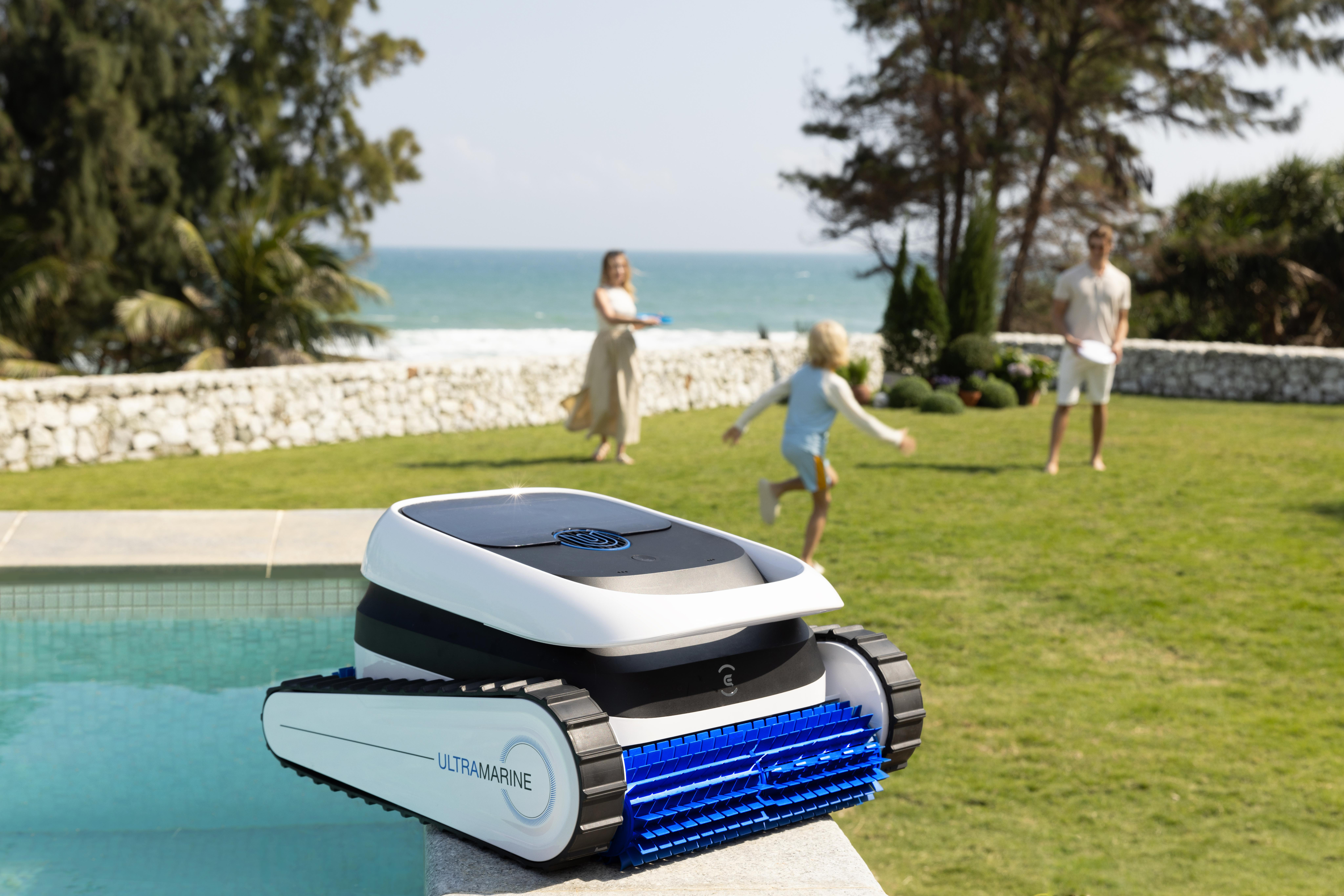 Ein weißer Poolreiniger mit blauen Bürsten steht am Beckenrand. Im Hintergrund spielen Menschen am Strand.