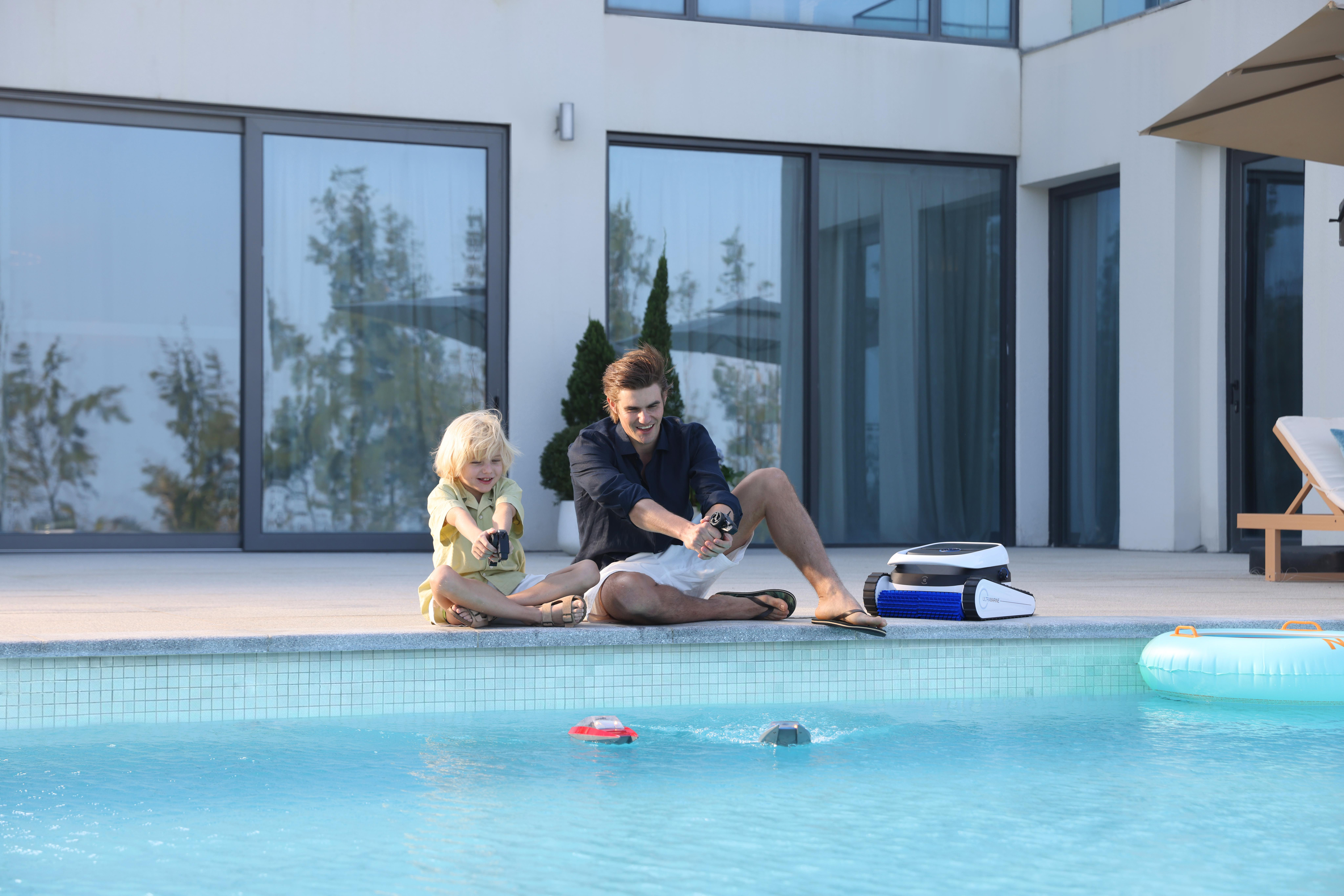 Mann und Kind sitzen am Pool und spielen mit ferngesteuerten Booten. Ein Poolreiniger steht daneben.