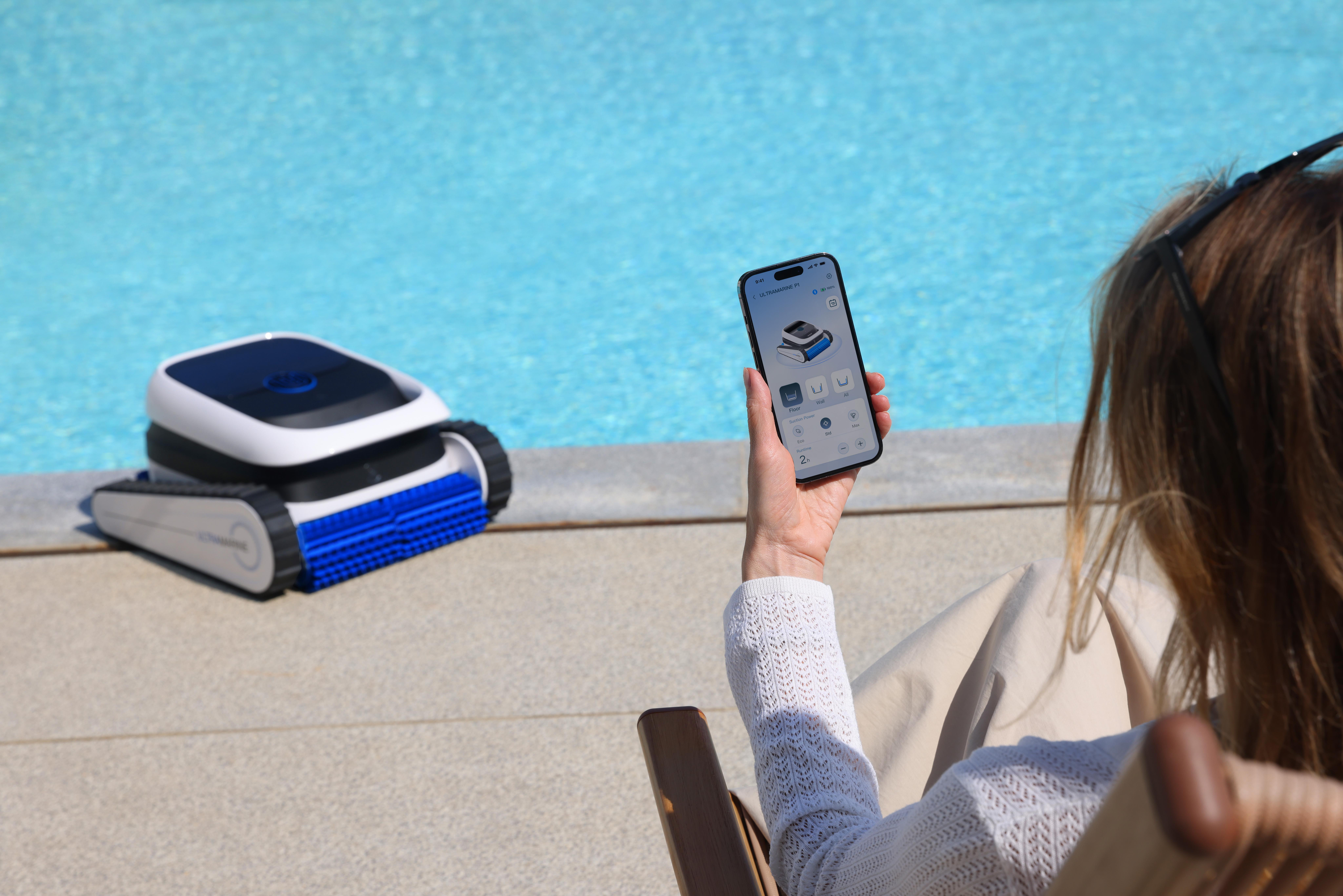 Ein Poolreiniger liegt am Beckenrand, eine Person hält ein Smartphone mit App.